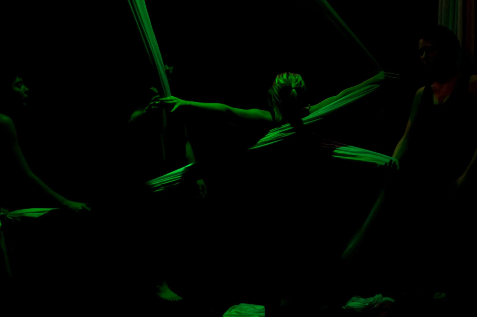 Three performers in black with glow-in-the-dark green fabric creating a dance or theatrical scene in dark lighting.
