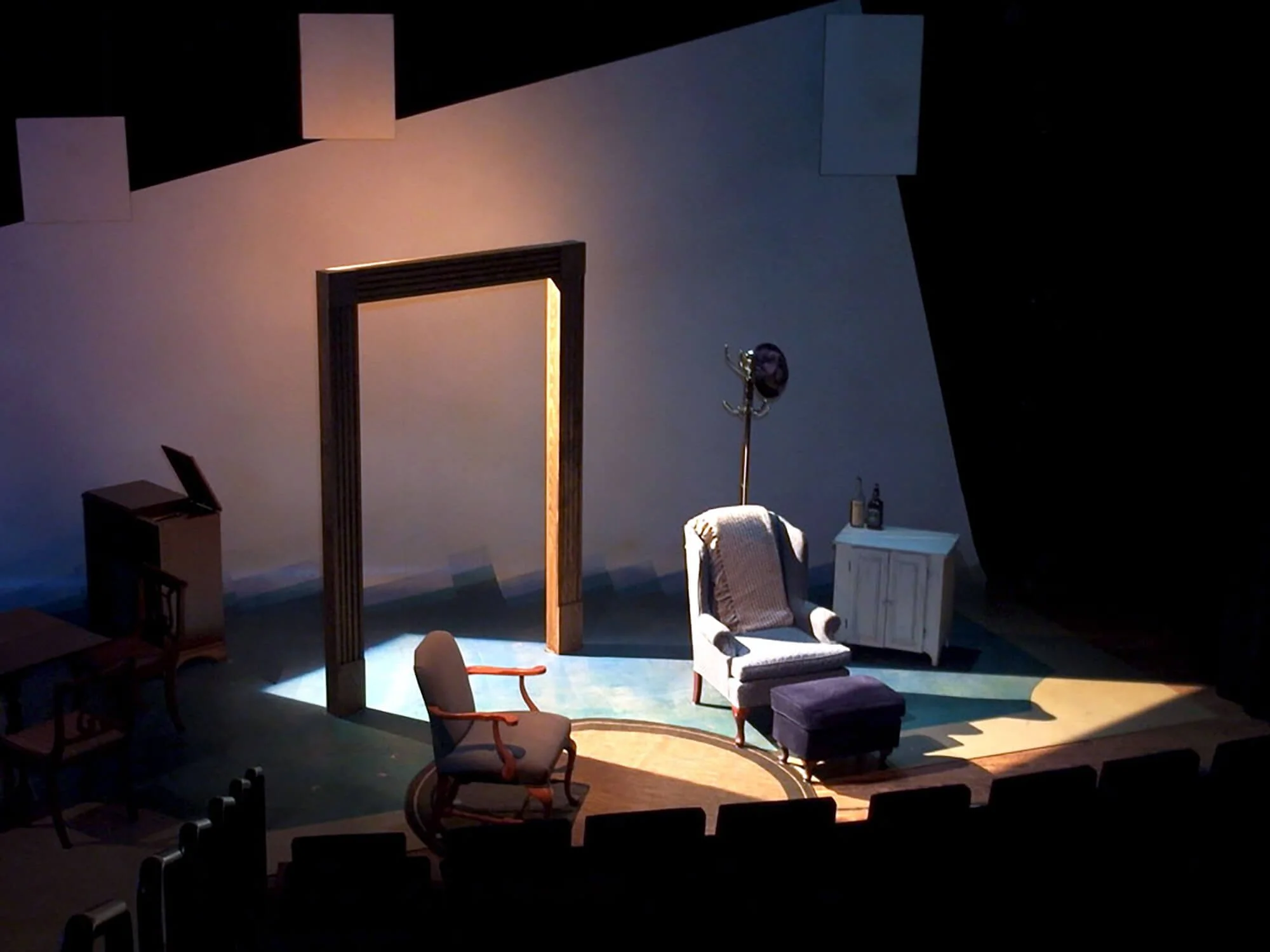 Stage set with a white armchair, a smaller purple ottoman, a beige curtain, a coat rack with a hat, small side table, and lamps, illuminated with stage lights.