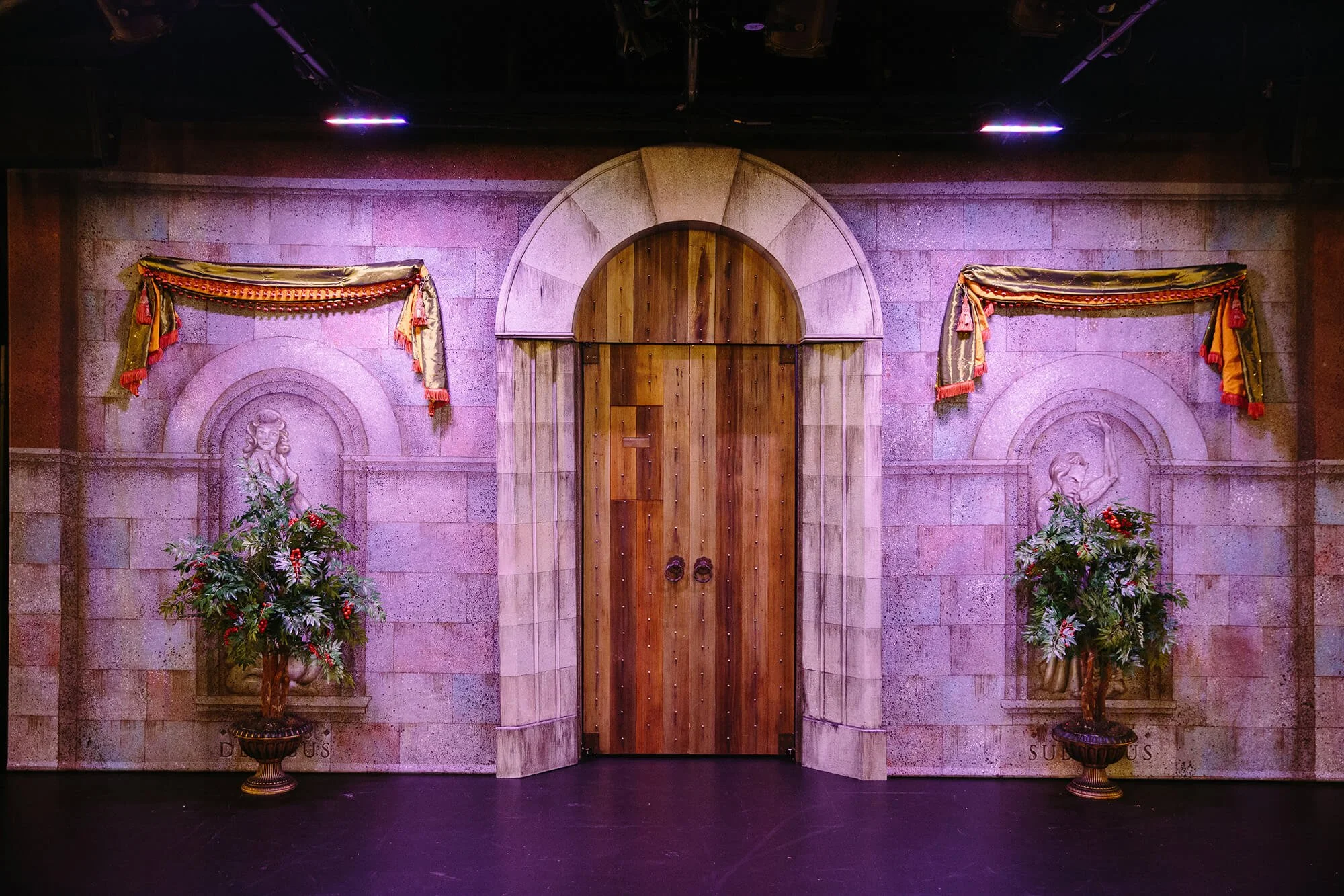 The image shows a stage with a wooden door centered, flanked by two statues of women in niches, adorned with floral arrangements. Ornate curtains hang above the statues, and the background features a textured stone wall with purple lighting.