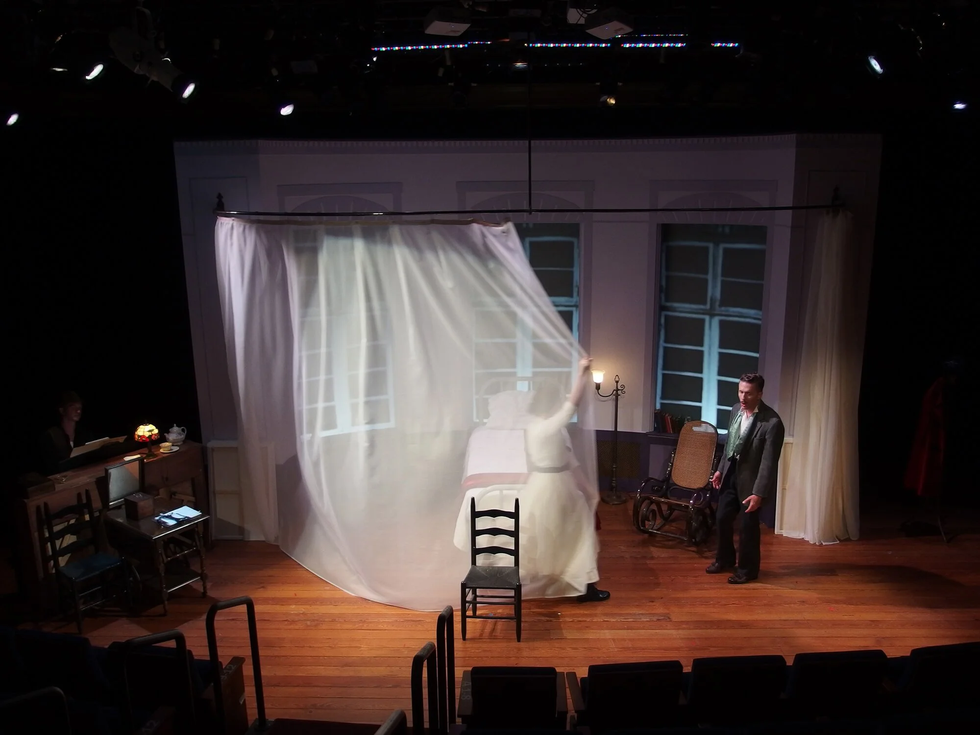A theater stage scene with a white bed, a woman in a white dress, and a man in a suit, with a curtain and windows in the background.