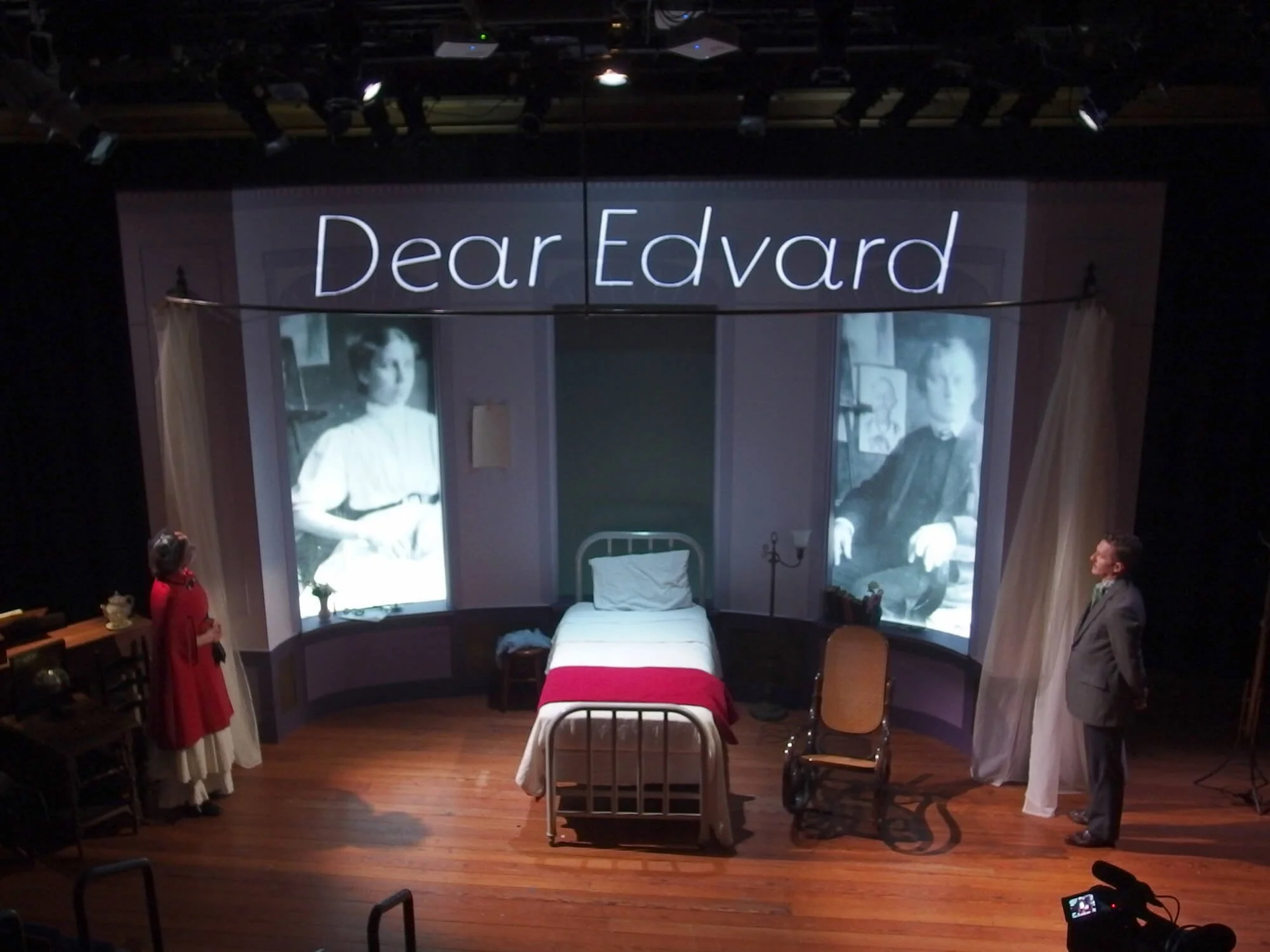 Stage scene with a bed in the center, two black-and-white projected photos of a woman on either side, a man in a gray suit on the right, and a woman in a red cloak on the left, with the words 'Dear Edvard' in neon above.