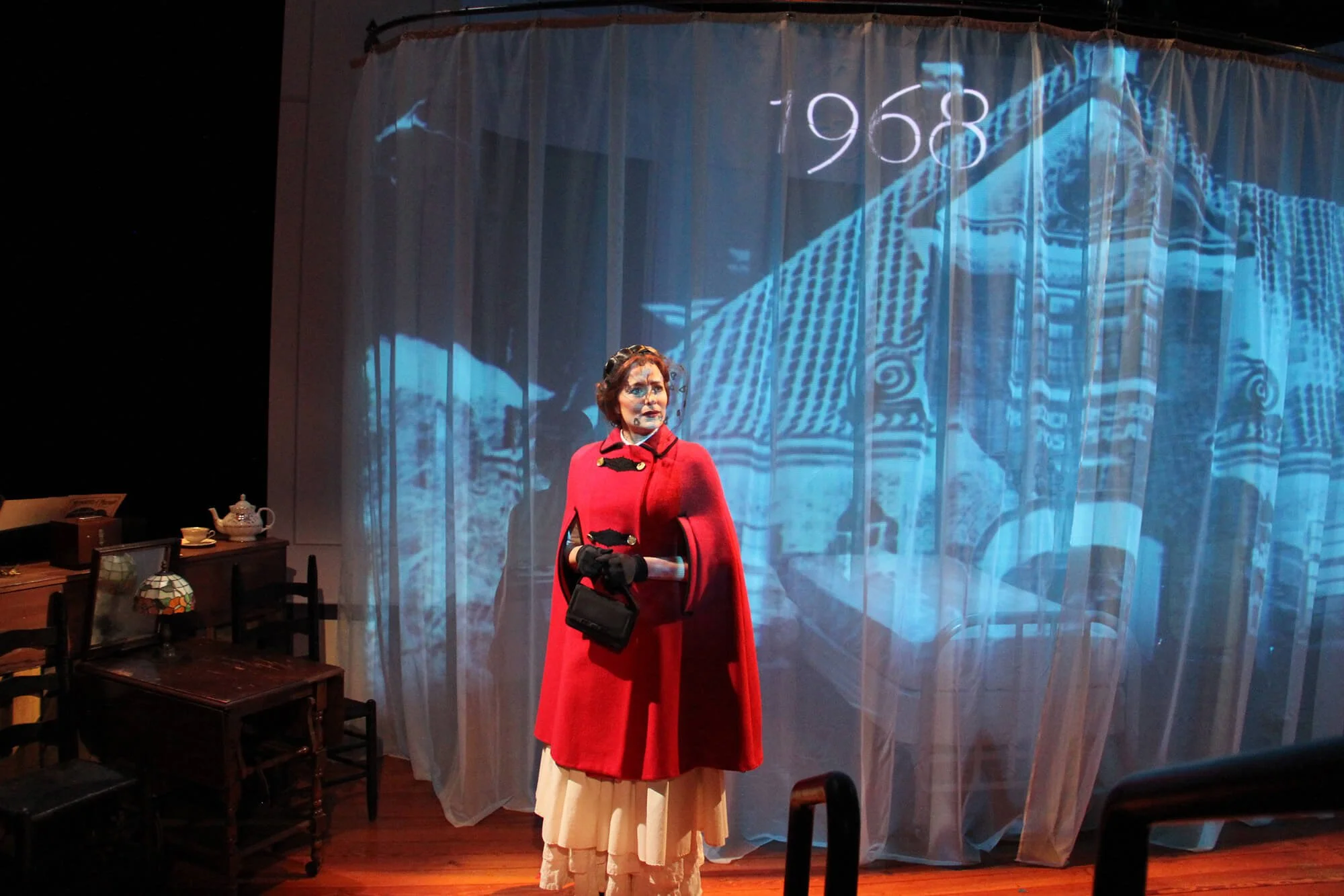A woman dressed in vintage clothing, wearing a red coat and black gloves, standing on a stage with a backdrop showing a blue-looking historic building with the year 1968 written on it.