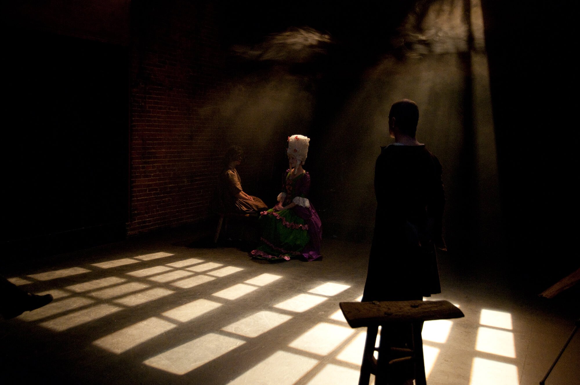 Dimly lit scene with four people, one woman seated and three standing, in period costumes, with a timestamped window casting shadows on the floor.