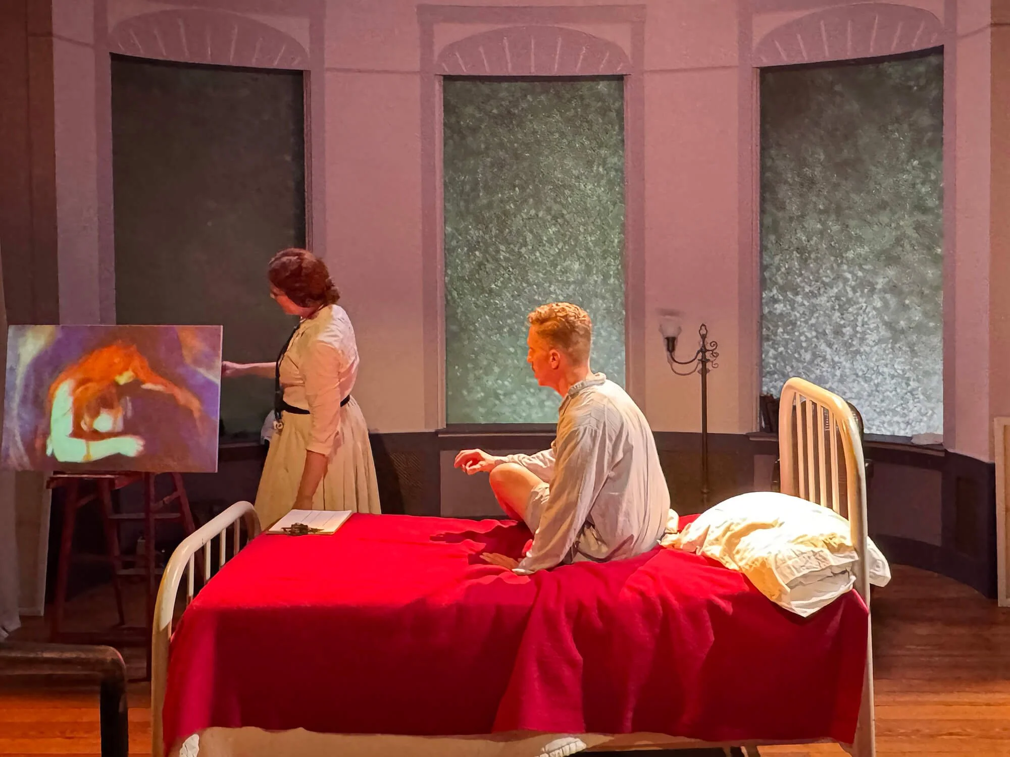 A scene with a man sitting on a bed with a red blanket, and a woman standing beside a painting in a room with large windows showing a rain or water scene.