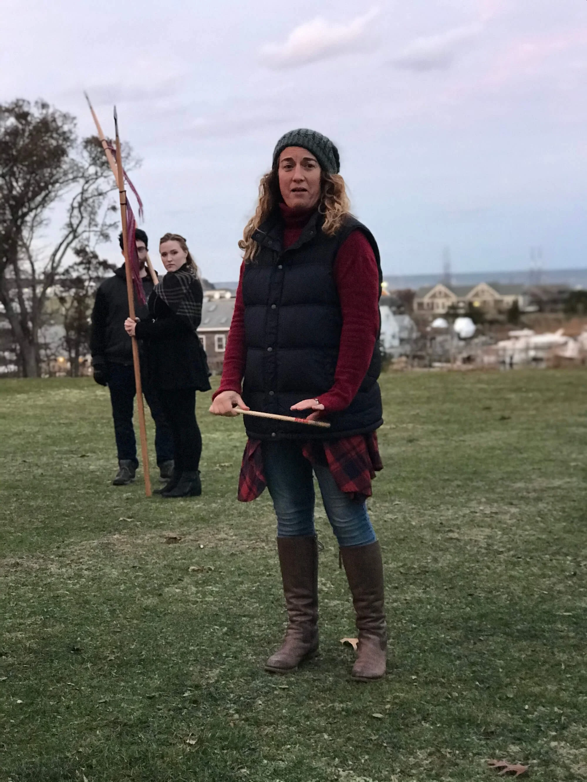 Woman in a gray knit hat, red sweater, black vest, jeans, and brown boots standing outdoors on a grassy area with a group of people in the background holding a flagpole, overcast sky, and distant houses.