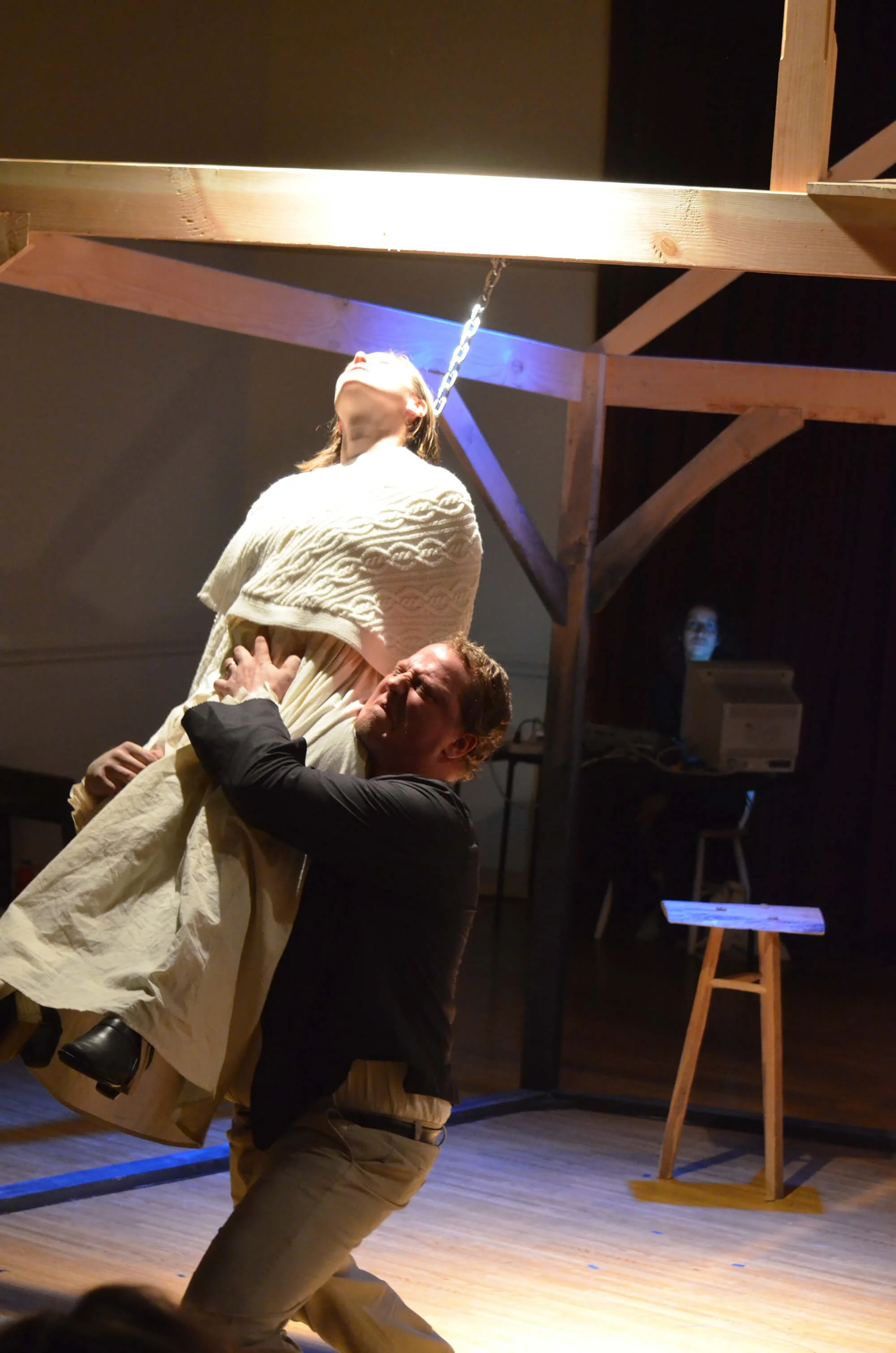 A theatrical scene with a man holding a woman who is suspended in the air, against a dark background with wooden beams and a stool. The woman is leaning back with her face directed upward, illuminated by dramatic lighting, and the man appears to be s