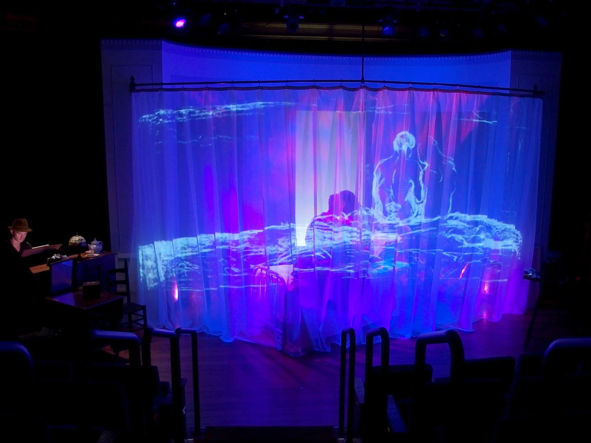 A theater stage with a semi-transparent curtain illuminated by colorful stage lights. Behind the curtain, a projection of a blue and purple abstract scene is visible, with a faint silhouette of a person sitting at a table and a glowing figure that ap