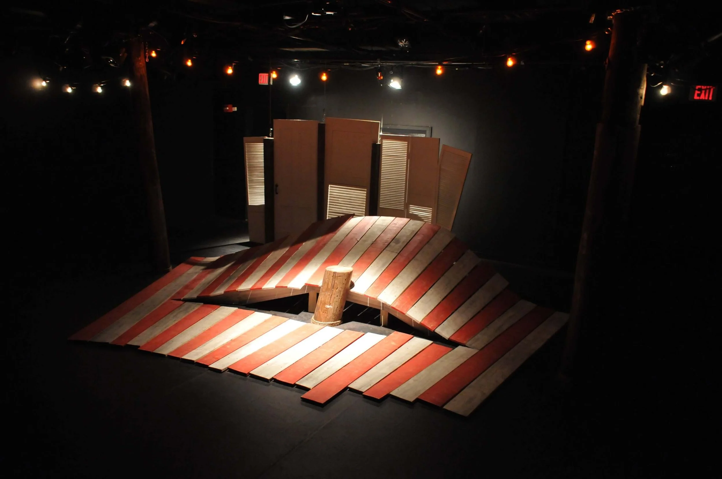 A theatrical set design resembling a collapsed fence or wall with a raised, curved platform on wooden planks, and a backdrop of multiple folding wooden shutters in a dark room with dim lighting.