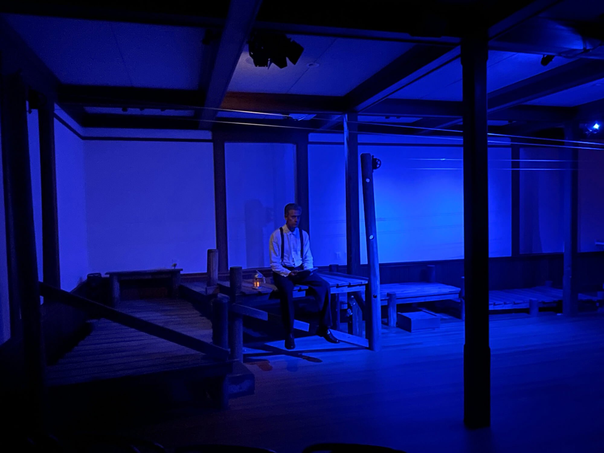 A man is sitting on a bench in a dimly lit room illuminated with blue light, with a wooden structure and some ropes or wires overhead.