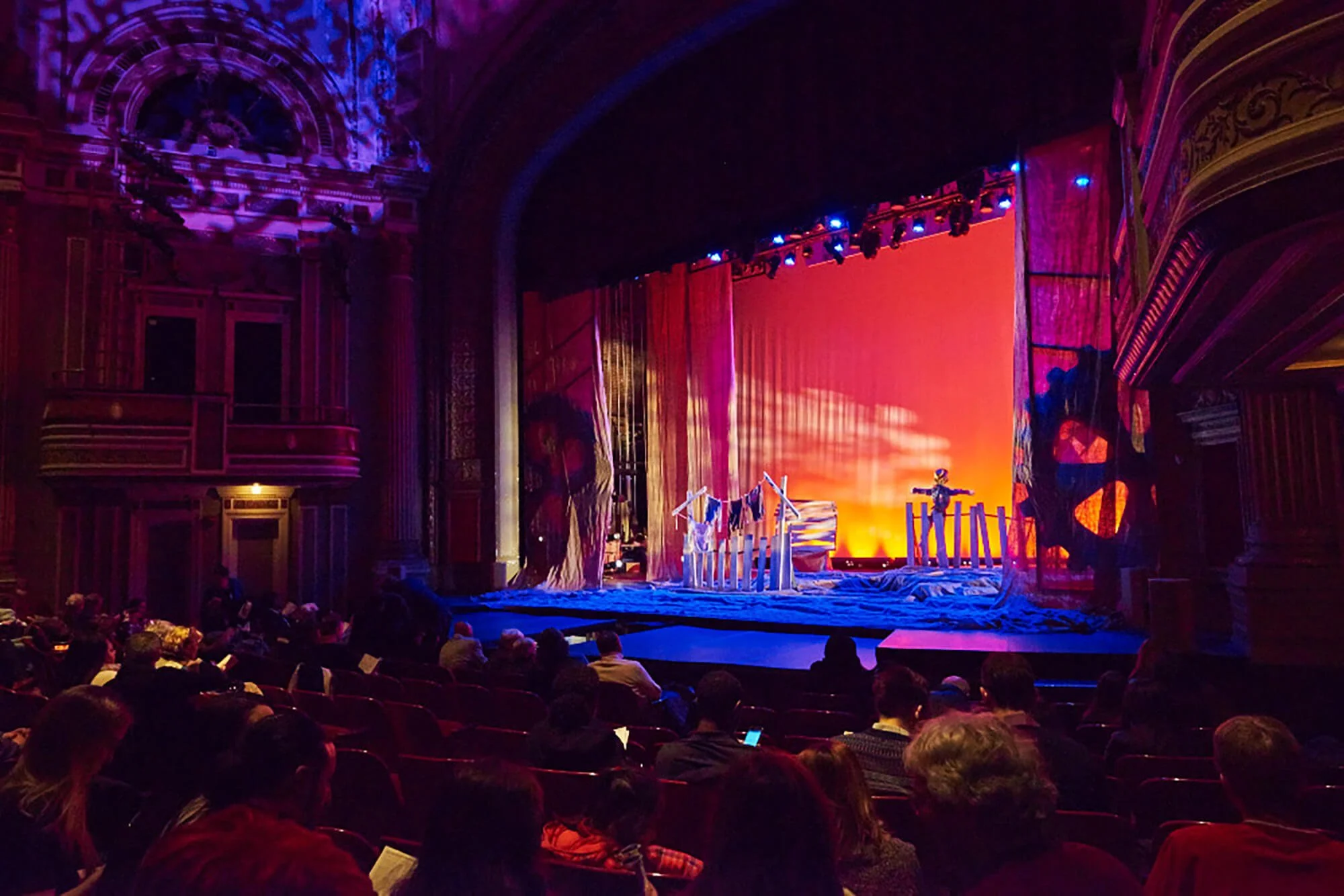 Theatrical stage set with colorful lighting, curtains, and props, with audience seated watching a performance.