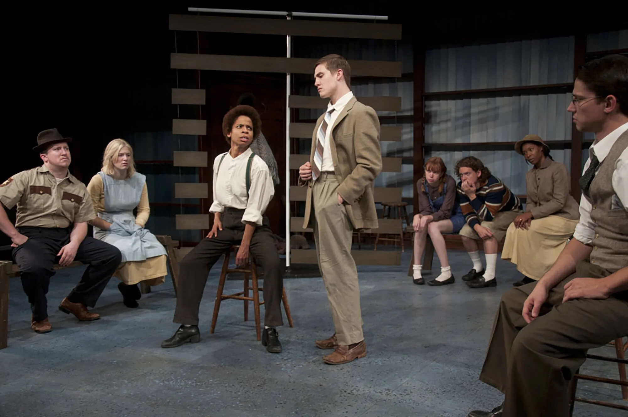 A theatrical performance with actors on stage, some seated and some standing, engaged in a dramatic scene.