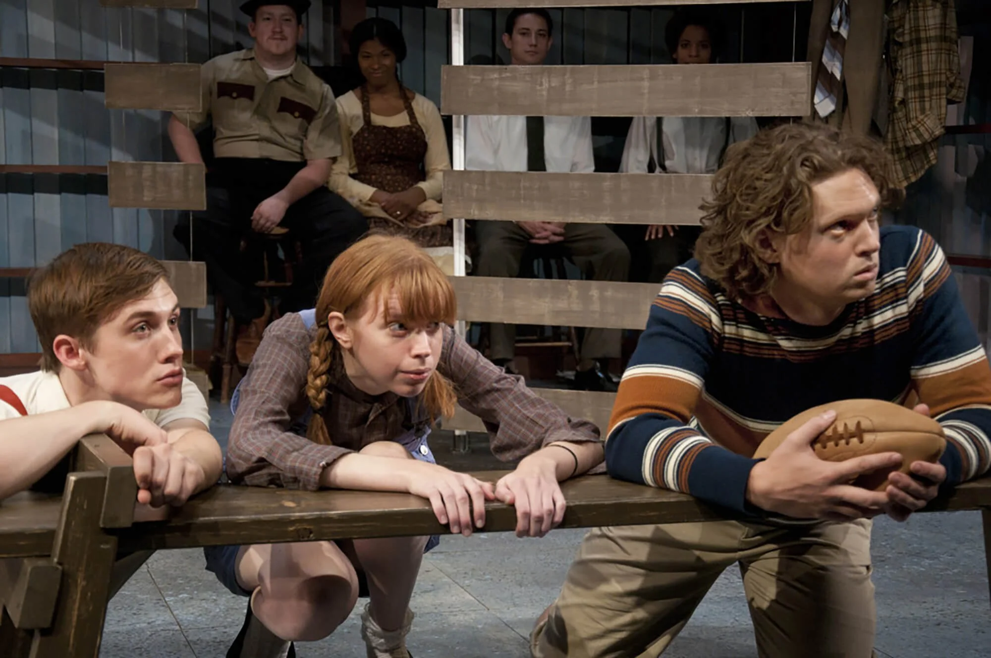 People sitting at a table, watching a man holding a football during a stage play or rehearsal.