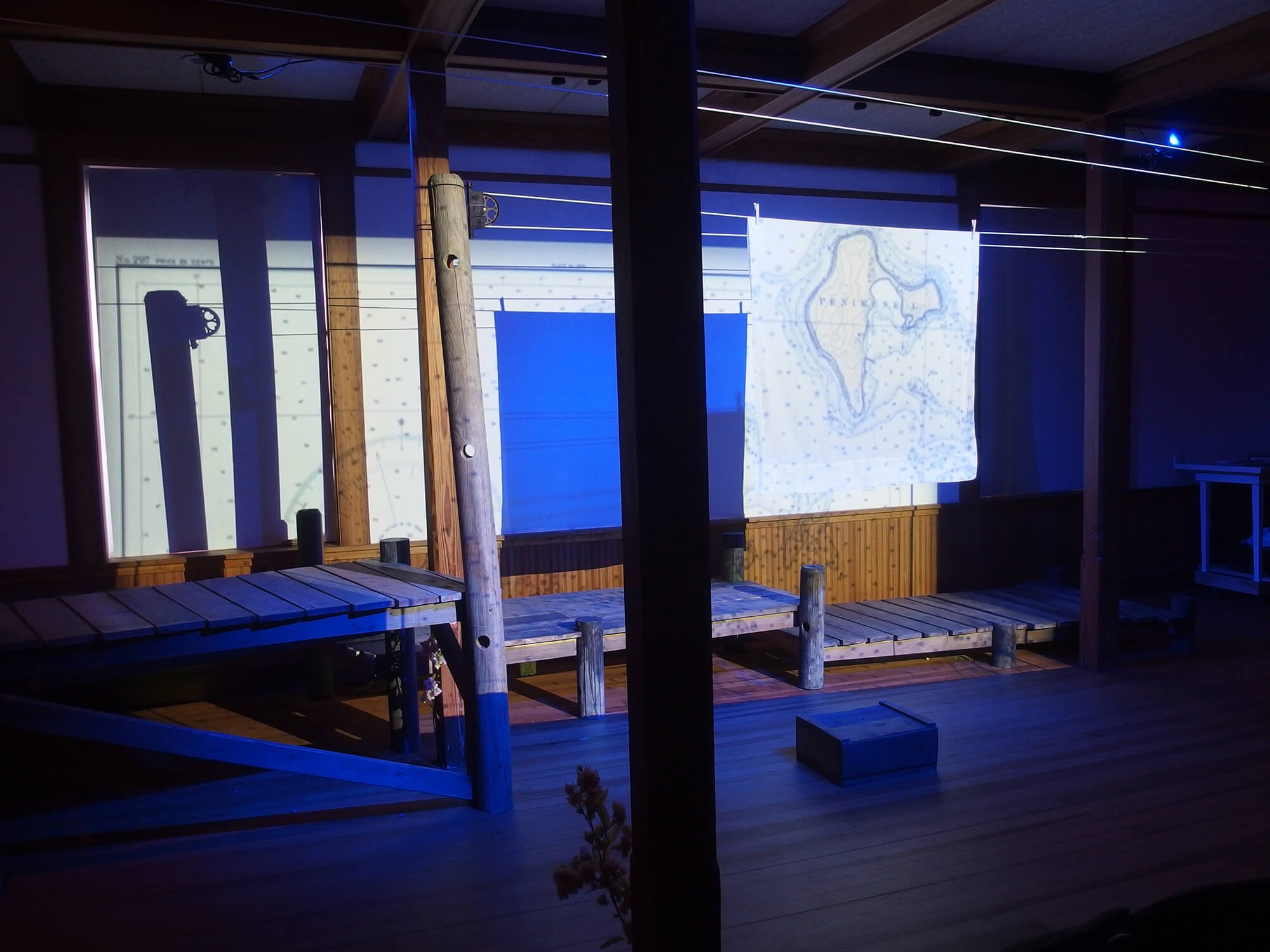 An indoor scene with a wooden deck, large windows, and a projection of a map of New Zealand on a piece of fabric hanging from a string. The space has dark wooden beams and minimal lighting.