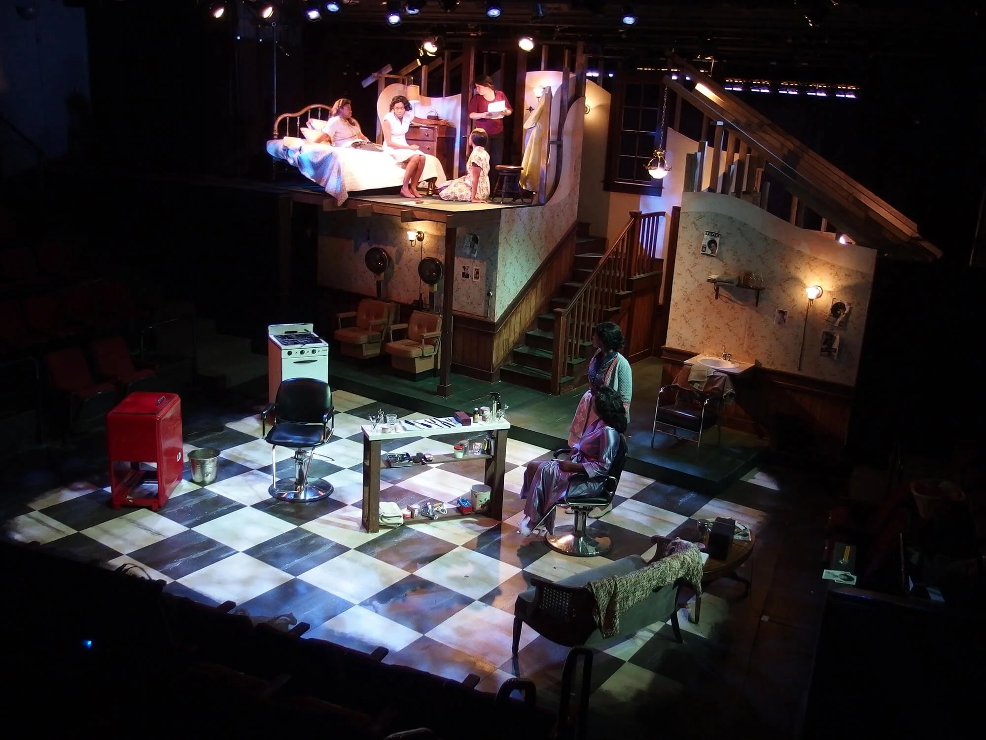 A theater stage set depicting a domestic scene with a woman lying in bed on the upper level, two women sitting and standing on the lower level, a kitchen stove, chairs, and various props, illuminated with warm lighting.
