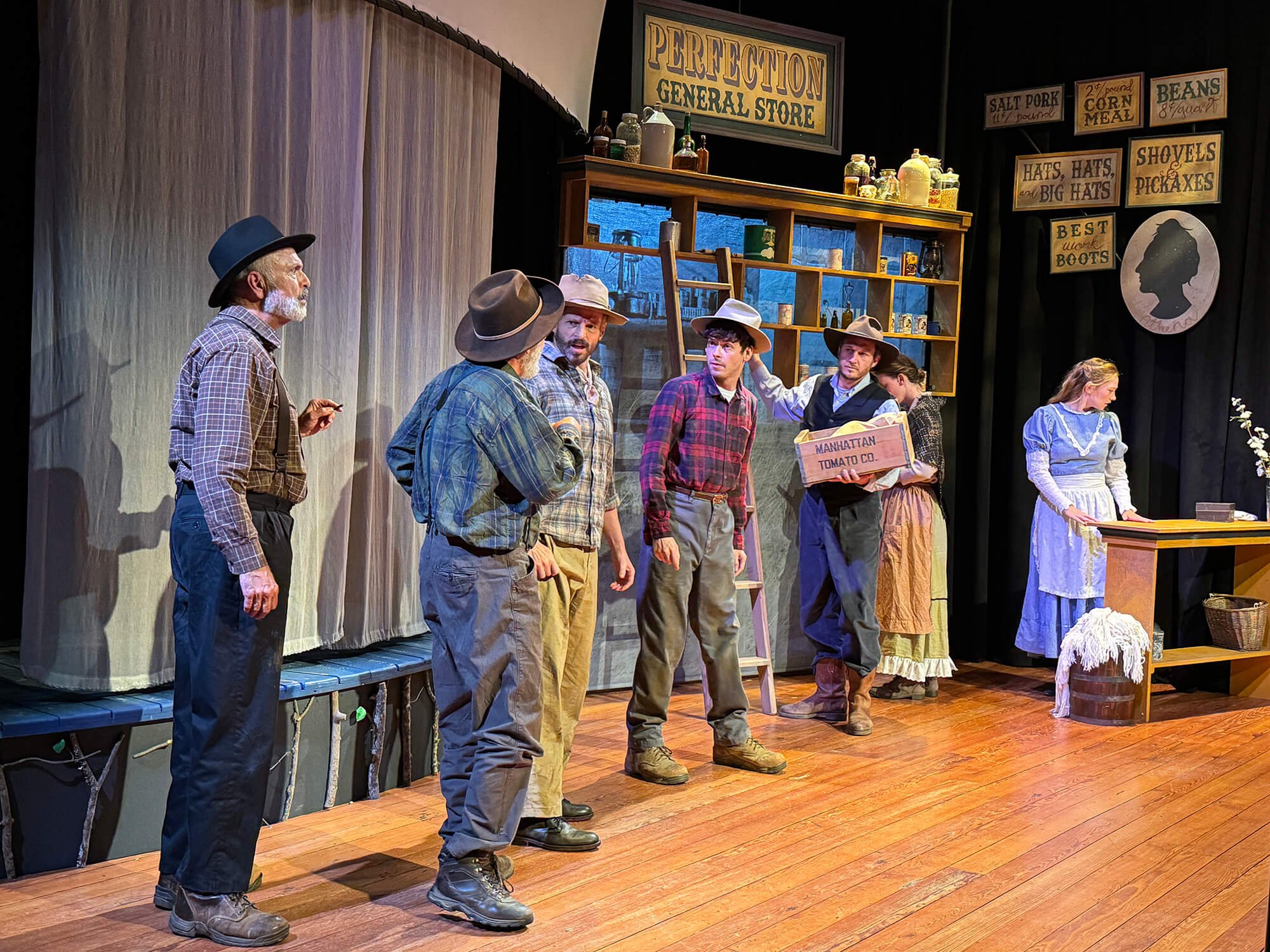A theatre stage depicting a scene with seven actors dressed in early 20th-century clothing, standing in front of a backdrop and props representing a general store. The setting includes signs and shelves with vintage items, with actors engaged in dial