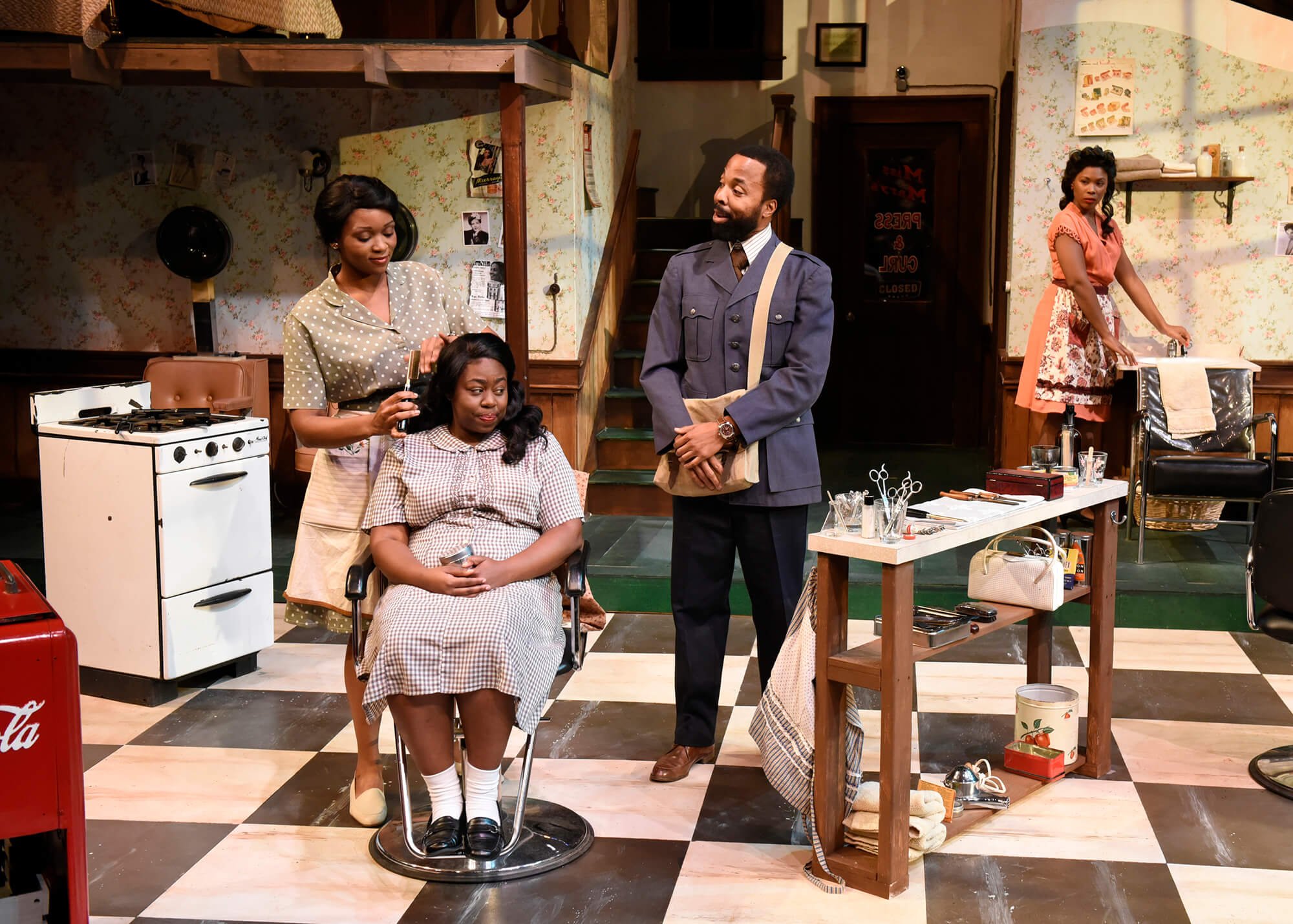 A woman is sitting in a barber chair while a second woman styles her hair. A man, dressed in a vintage suit, stands nearby holding a shopping bag, observing their actions. In the background, another woman is working at a sink. The setting appears to 