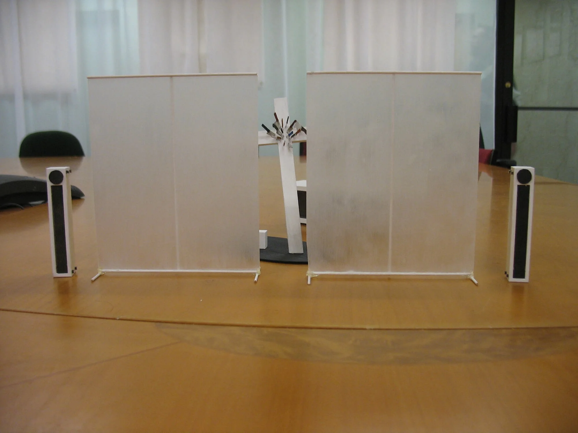 A miniature model of a scene with two large transparent panels, two tall speakers, a small stage with a man in a red shirt, and an abstract sculpture in the background.