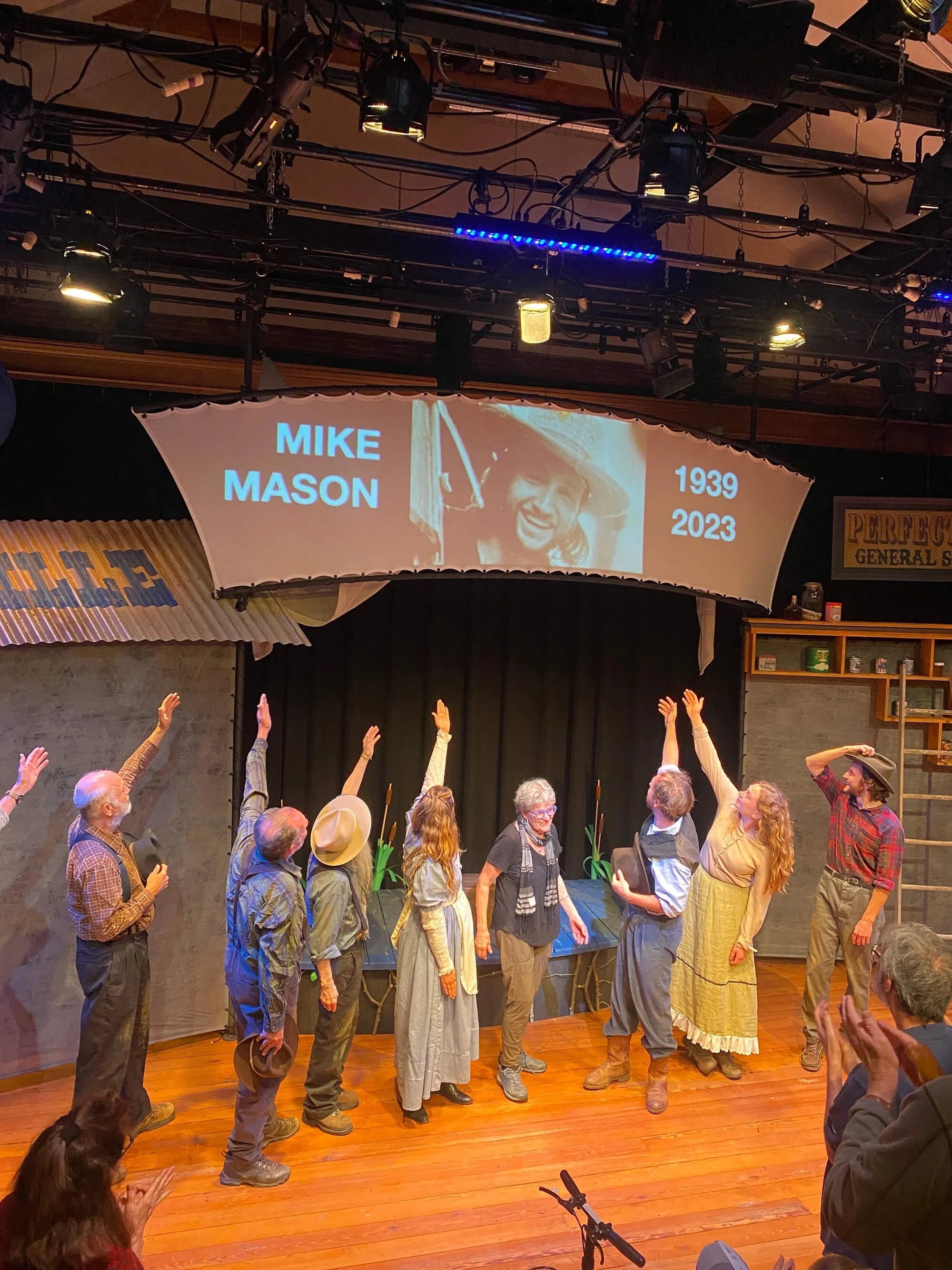 Stage scene with actors in period costumes, some raising their hands, in front of a black curtain. Above, a projected tribute to Mike Mason, featuring his name, photo, and the years 1939 to 2023. Audience members are applauding.