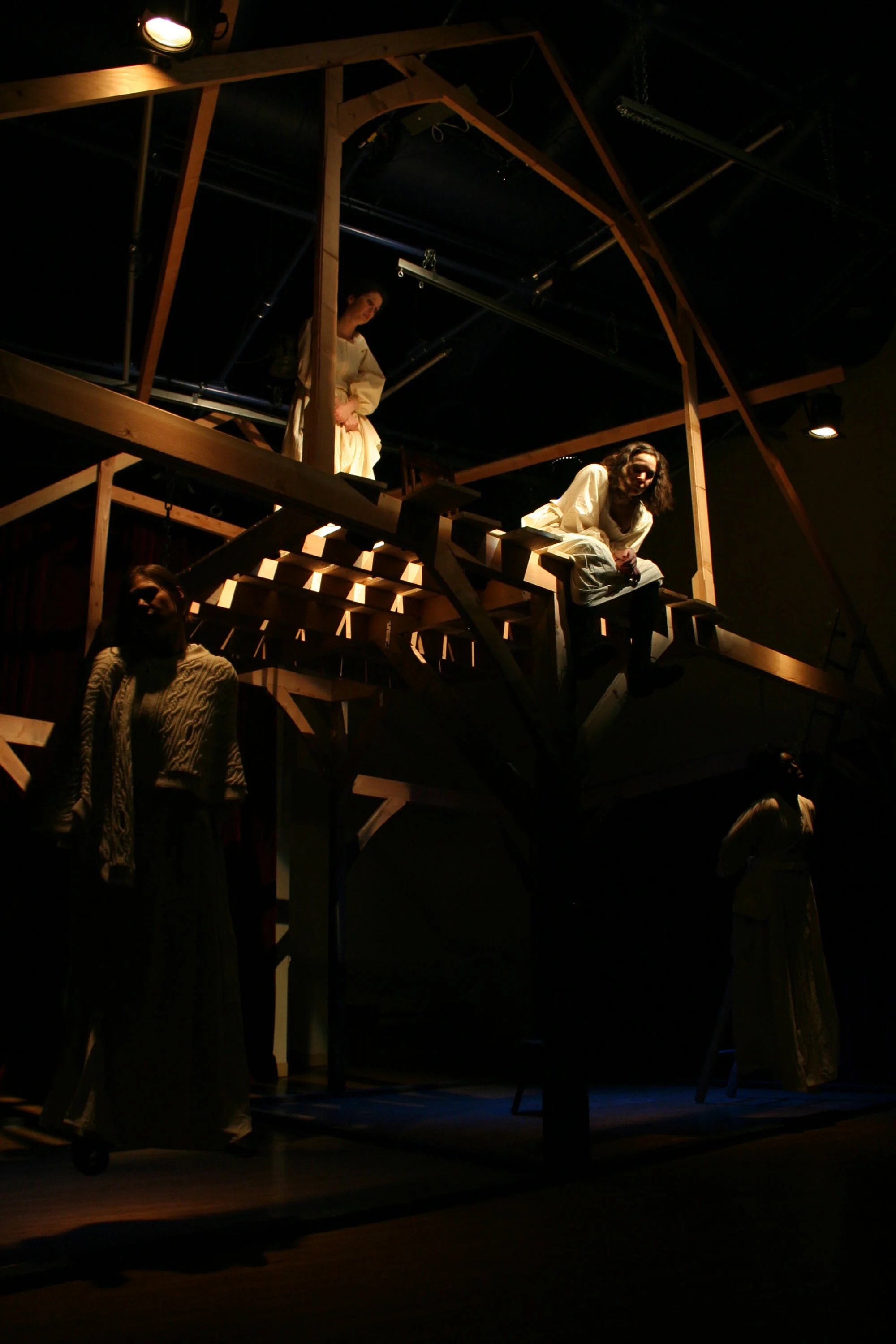 Theatrical stage set with a wooden treehouse structure, three actors in period costume, dim lighting with some spotlights, and a dark background.