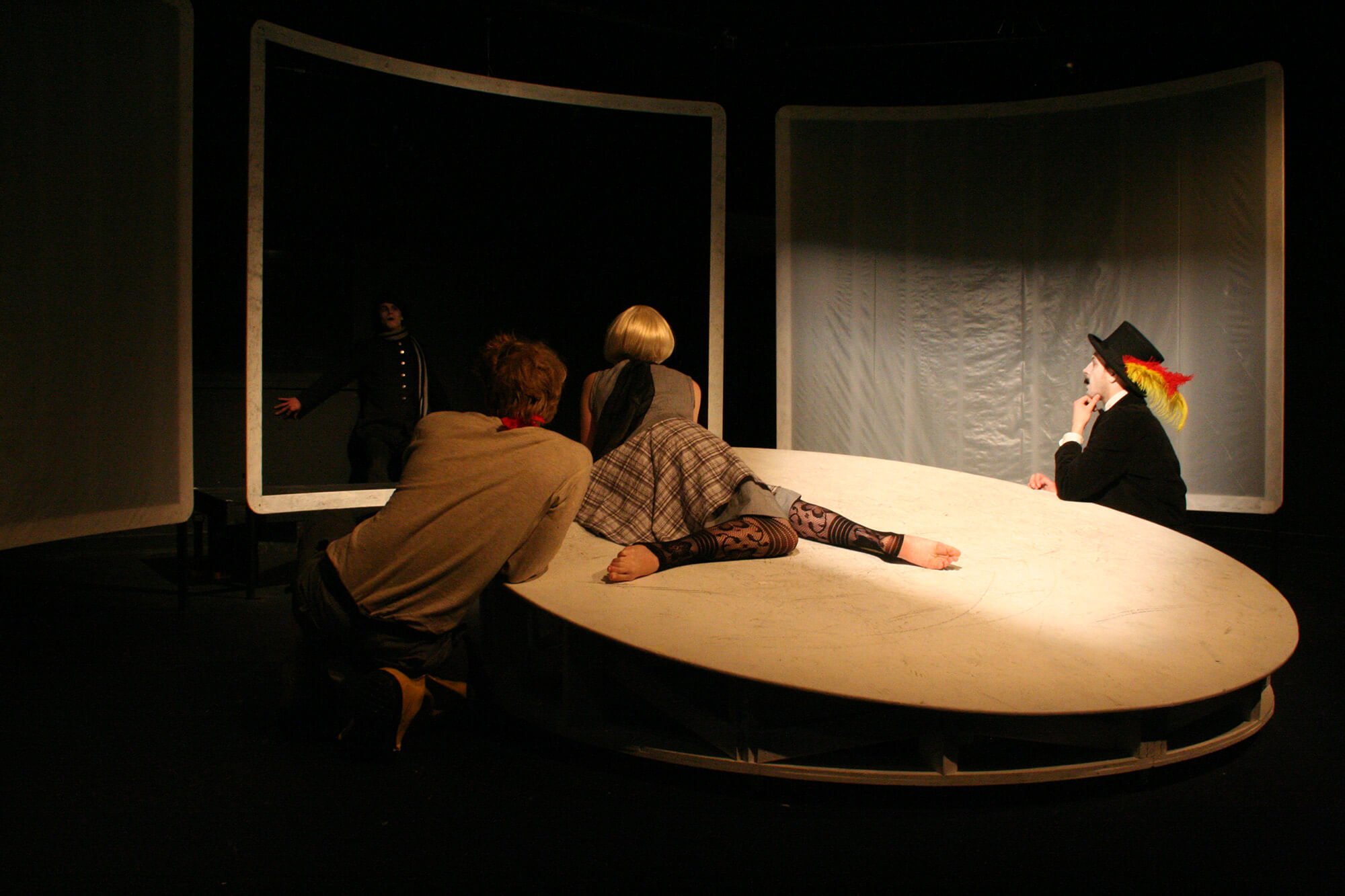 The image is of a theater scene with four actors. One actor is dressed in black with a feathered hat, sitting at a round table, deep in thought. Two actors are leaning over the table, one with red hair and the other with blonde hair, both observing t