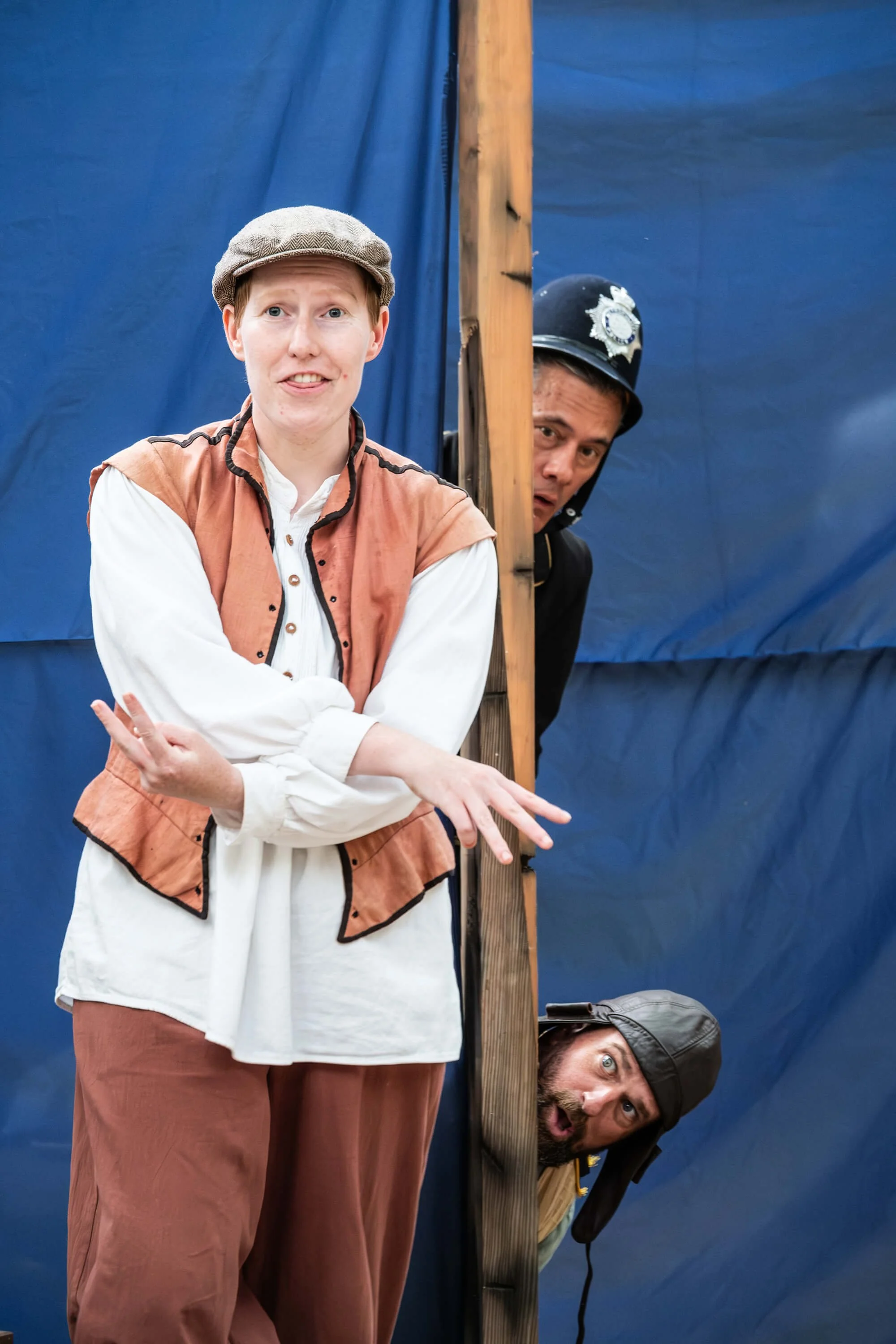 Three actors on stage in costume, with two peeking from behind a wooden partition and one standing in front, wearing costumes resembling early 20th-century military or police uniforms and casual clothing, with a blue backdrop.