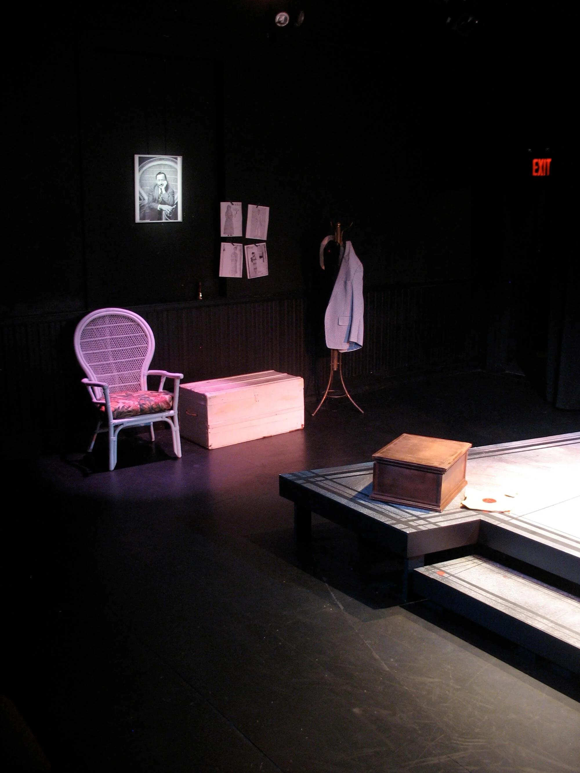 Stage with a white chair, a wooden box, a coat stand with a jacket, framed photo and drawings on the wall, and a red exit sign in the background.