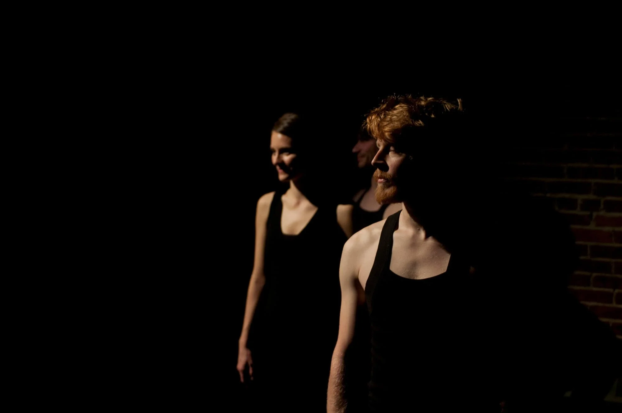 Three performers in black outfits standing in a row with dramatic lighting, against a dark background.