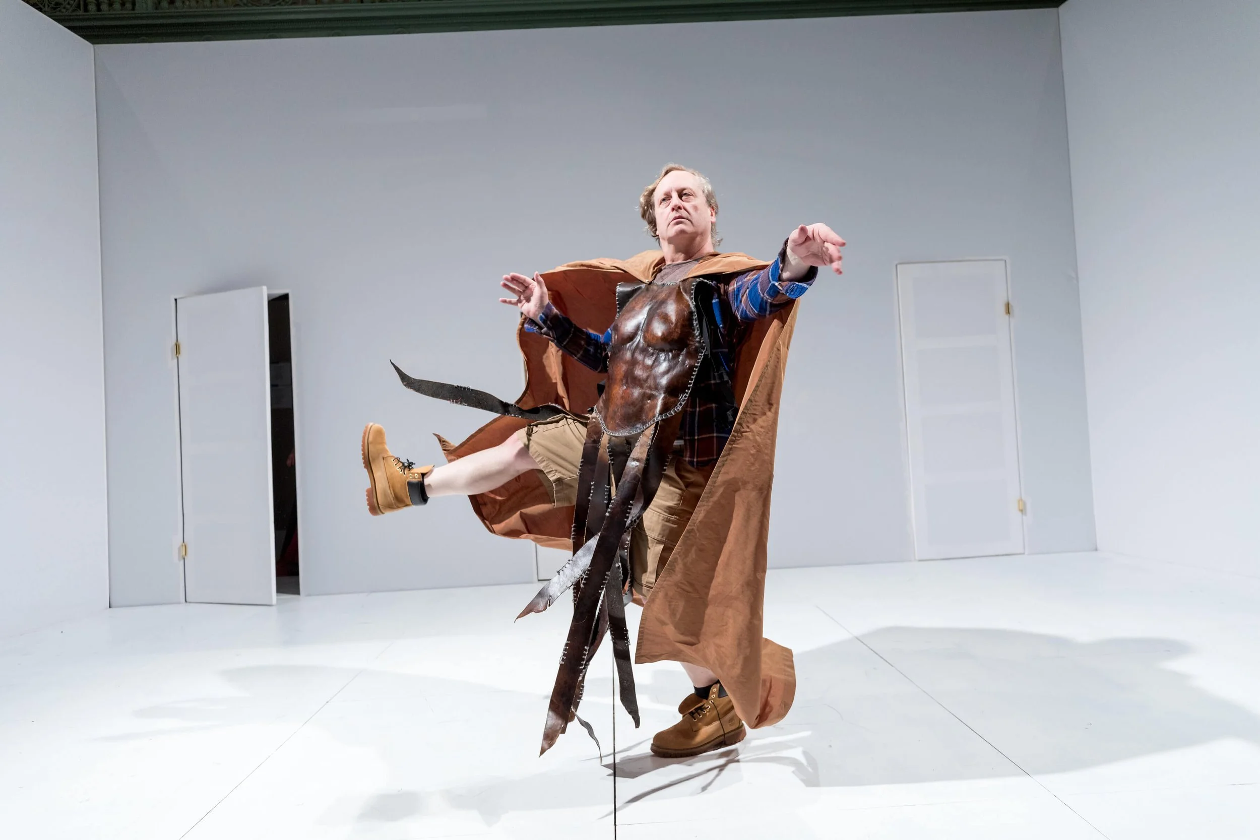 A man in costume performing a dance or pose in a minimalist white room, wearing a brown cloak, shorts, boots, and a chest piece with leather straps and feathers.