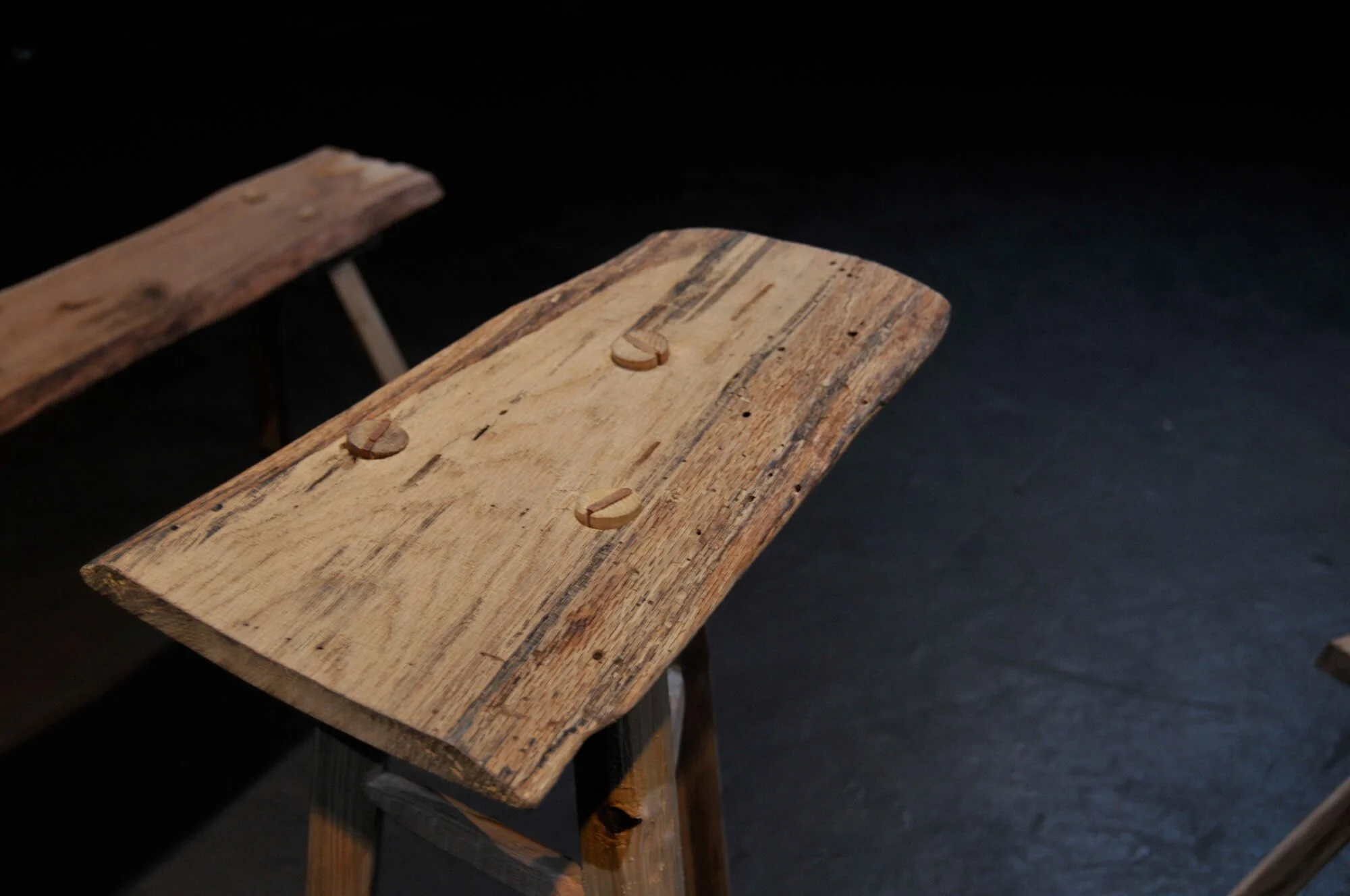 Close-up of a rustic wooden bench made from a single piece of aged wood with visible natural grain and small wooden dowels, set against a dark background.