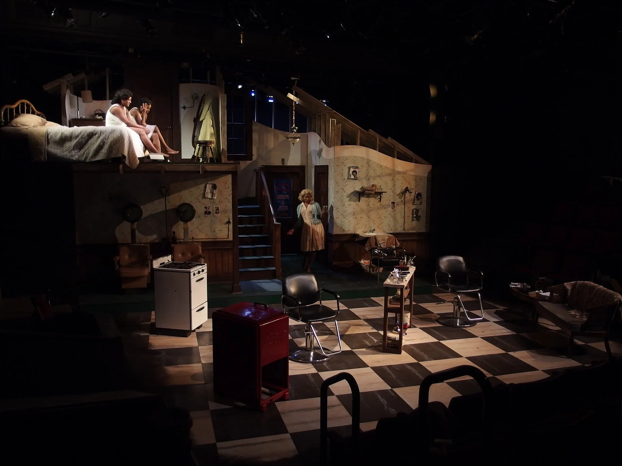 Stage set resembling a vintage domestic scene with a kitchen and a second-floor bedroom, featuring actors, furniture, and props for a theatrical production.