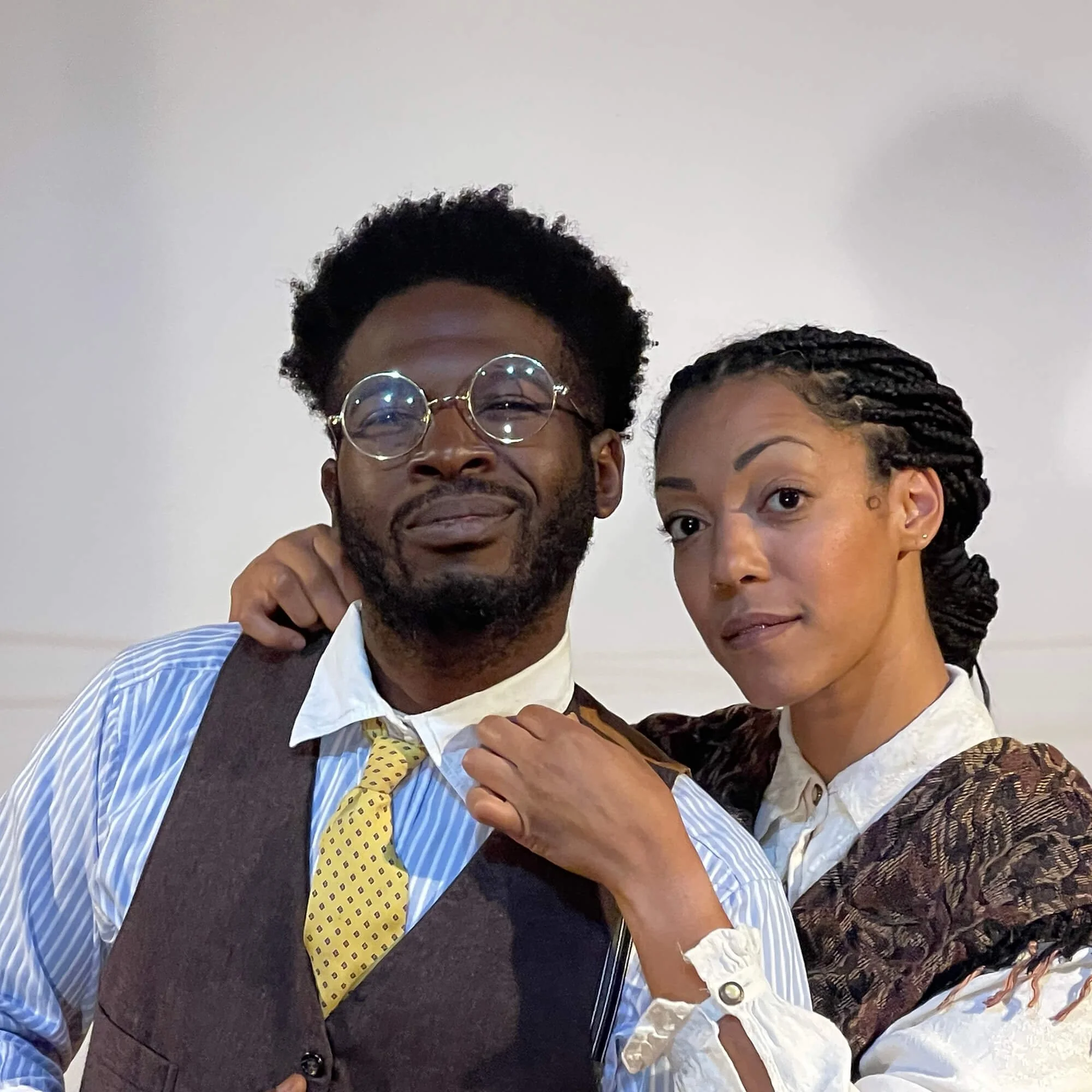 A man and woman posing together indoors, with the woman resting her hand on the man’s shoulder. The man wears large round glasses, a yellow tie with blue dots, a white shirt with blue stripes, and a brown vest. The woman has dark braided hair, well-d