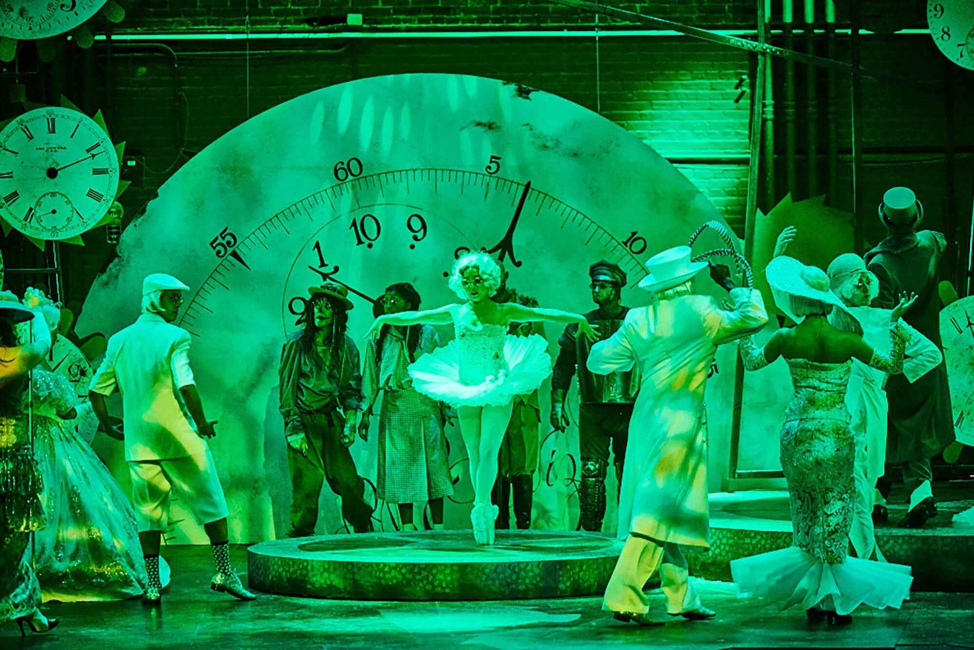 A theatrical stage with performers in vintage costumes, a large clock backdrop, and props, including clocks and a platform, suggesting a Tim Burton style production.