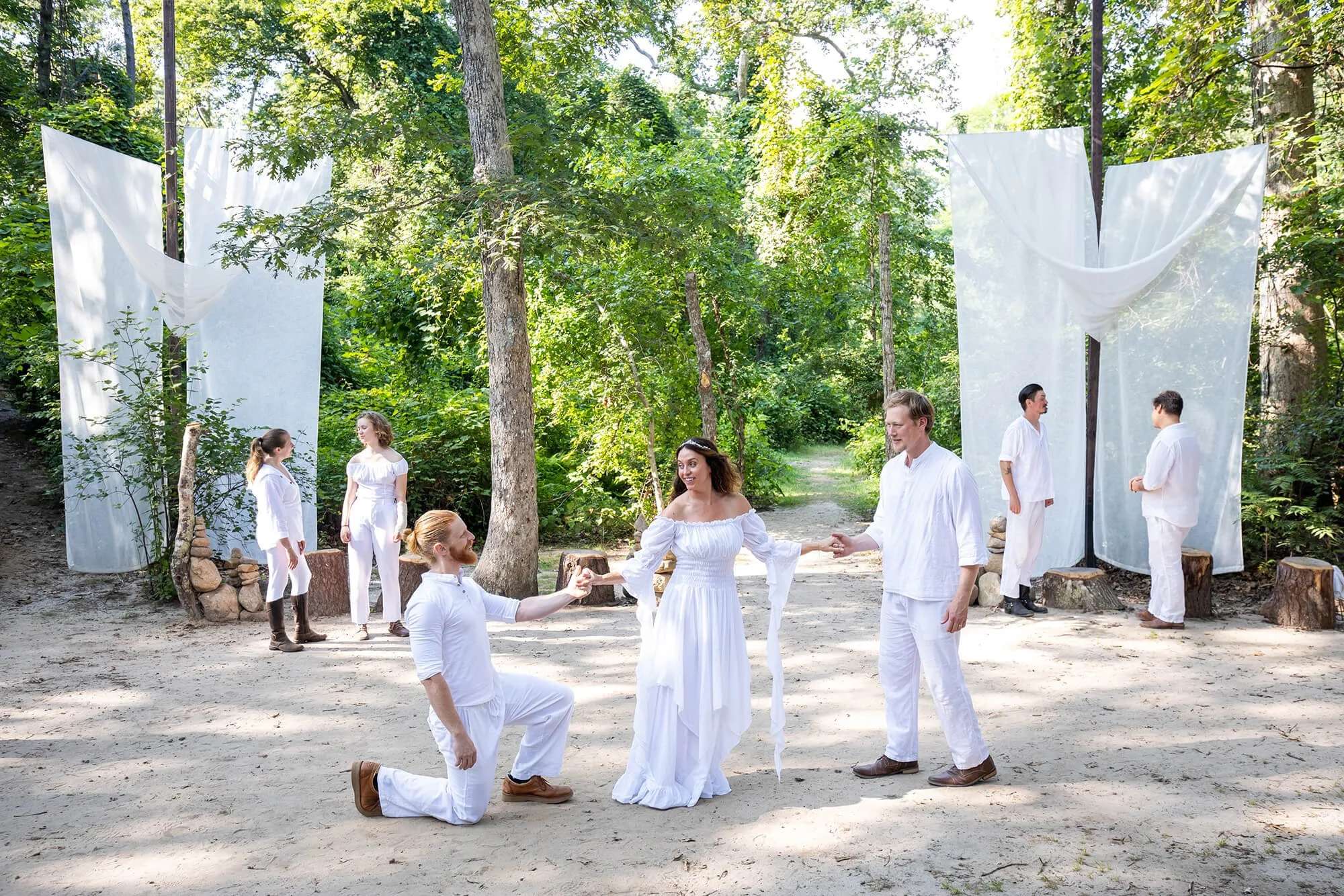 People dressed in white clothing participating in a wedding ceremony outdoors in a wooded area with trees and greenery, with some standing and others kneeling or holding hands.