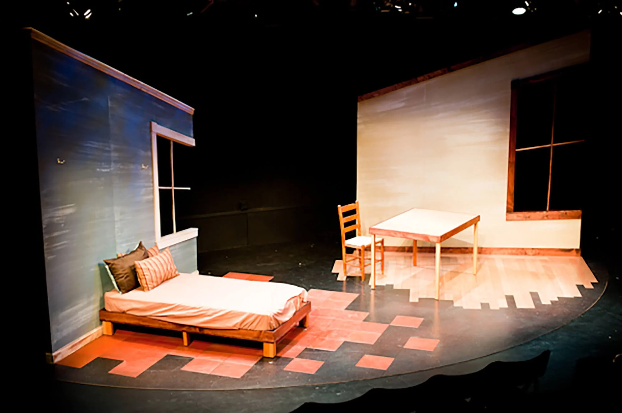 Stage set designed to look like a bedroom with a bed, pillows, a window, a table, and a chair, with a backdrop of blue and beige walls.