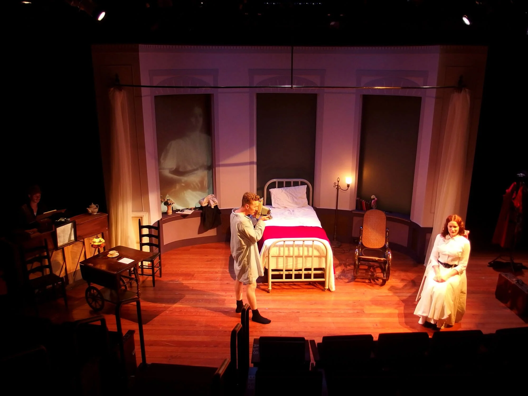 Stage scene from a play with three actors. One is standing and talking, one is sitting on a chair, and the third is sitting on a bed. The set depicts a bedroom with a bed, a wheelchair, a lamp, and other furniture.