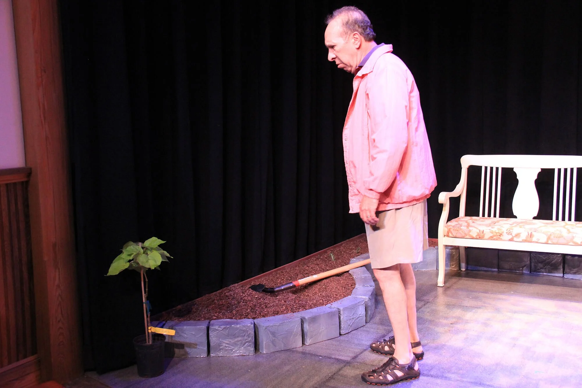 Man standing on stage with a gardening shovel, a small potted plant, and a garden bed with soil, in front of a black curtain, with a white bench on the right.