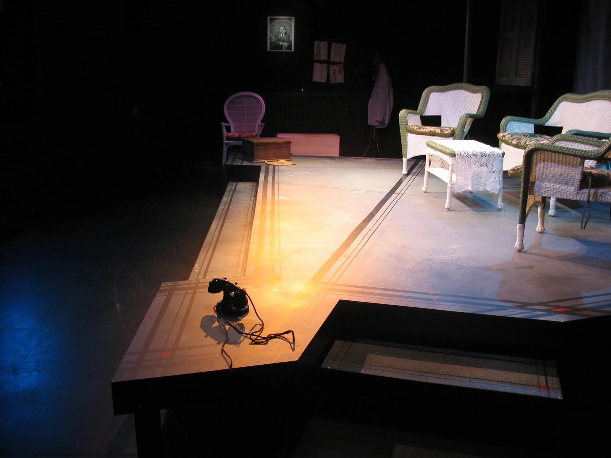Empty theater stage with vintage furniture including armchairs, a small table, a trunk, and a wooden chest, dimly lit with a spotlight, and a photograph hanging on the back wall.