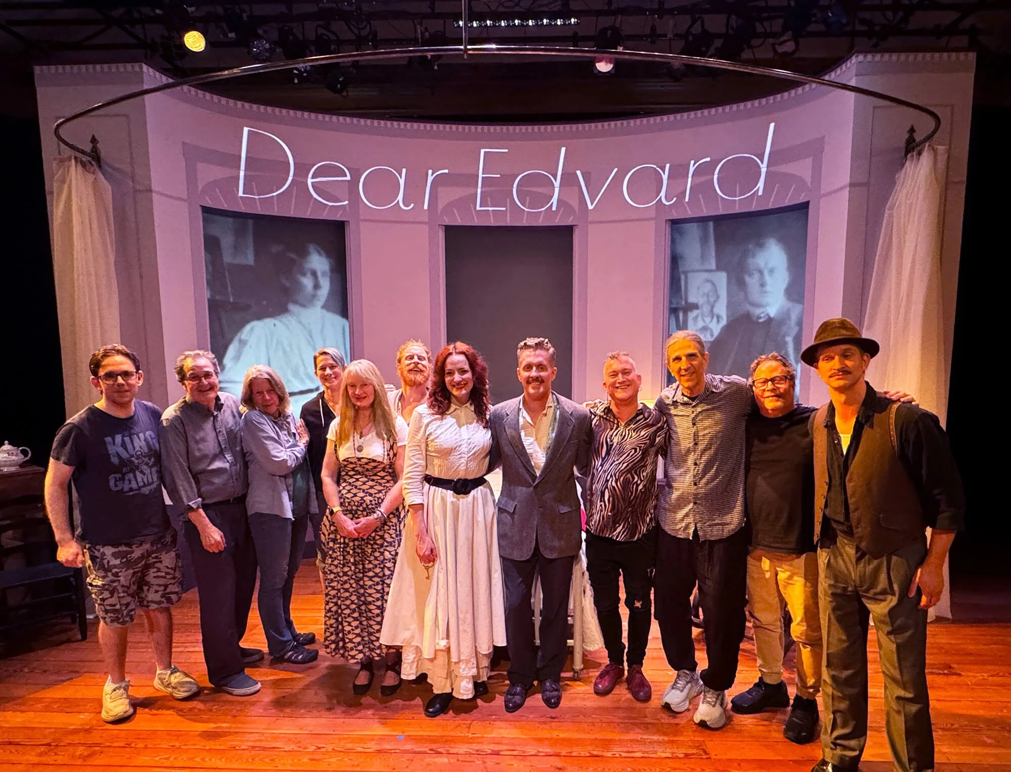 A group of people standing on a stage in front of a pink backdrop with the words "Dear Edvard" and black-and-white photos of a woman and a man. The group includes men and women dressed in casual and vintage-style clothing, and appears to be a cast or