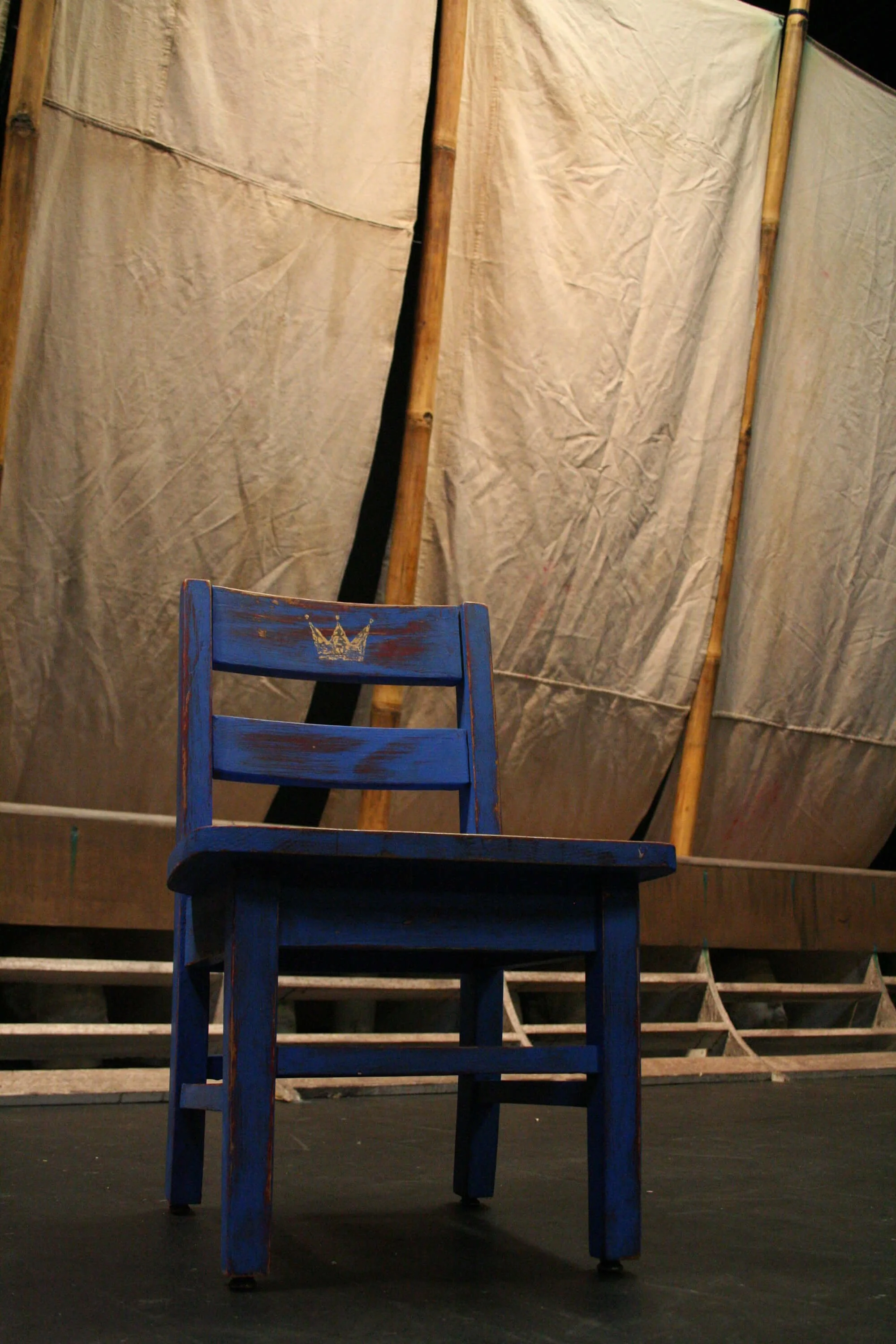 A blue wooden chair with a small gold crown symbol on the backrest, positioned on a stage with a beige fabric backdrop and wooden support beams.