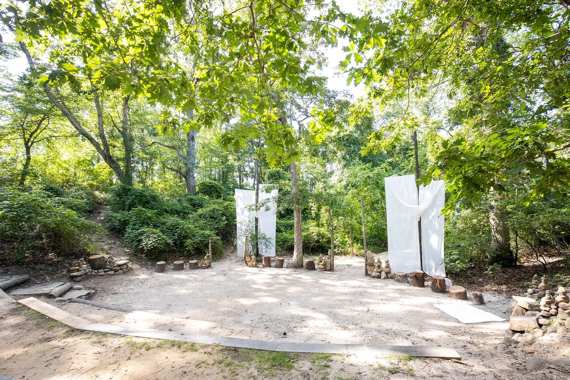 Outdoor wooded area with white fabric drapes hanging between trees, arranged in a semi-circle with small stacked rocks at the base of each drape, on a dirt ground.