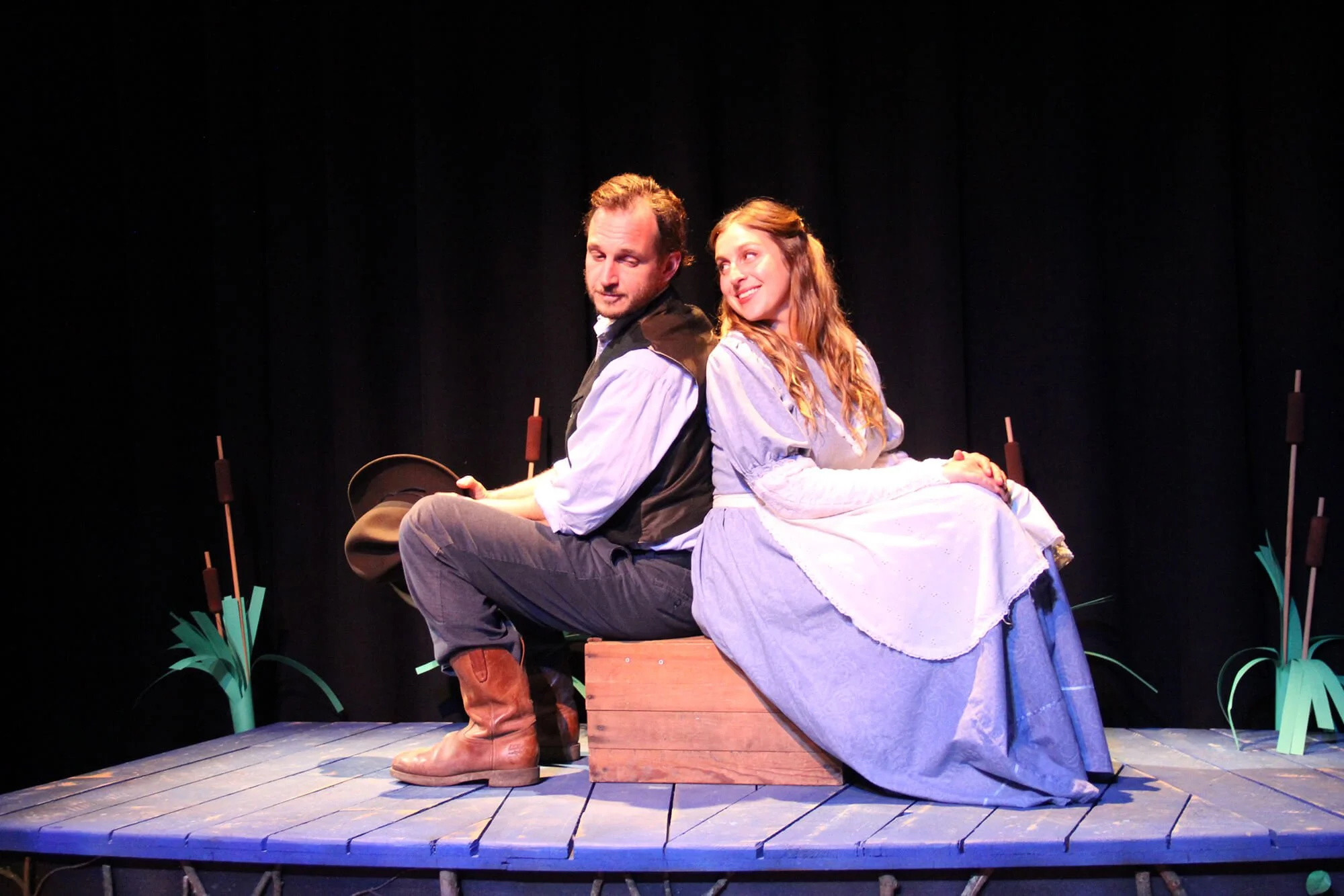 Actors on stage performing in costume, sitting back to back on a wooden platform, with a dark background and stage props resembling plants.