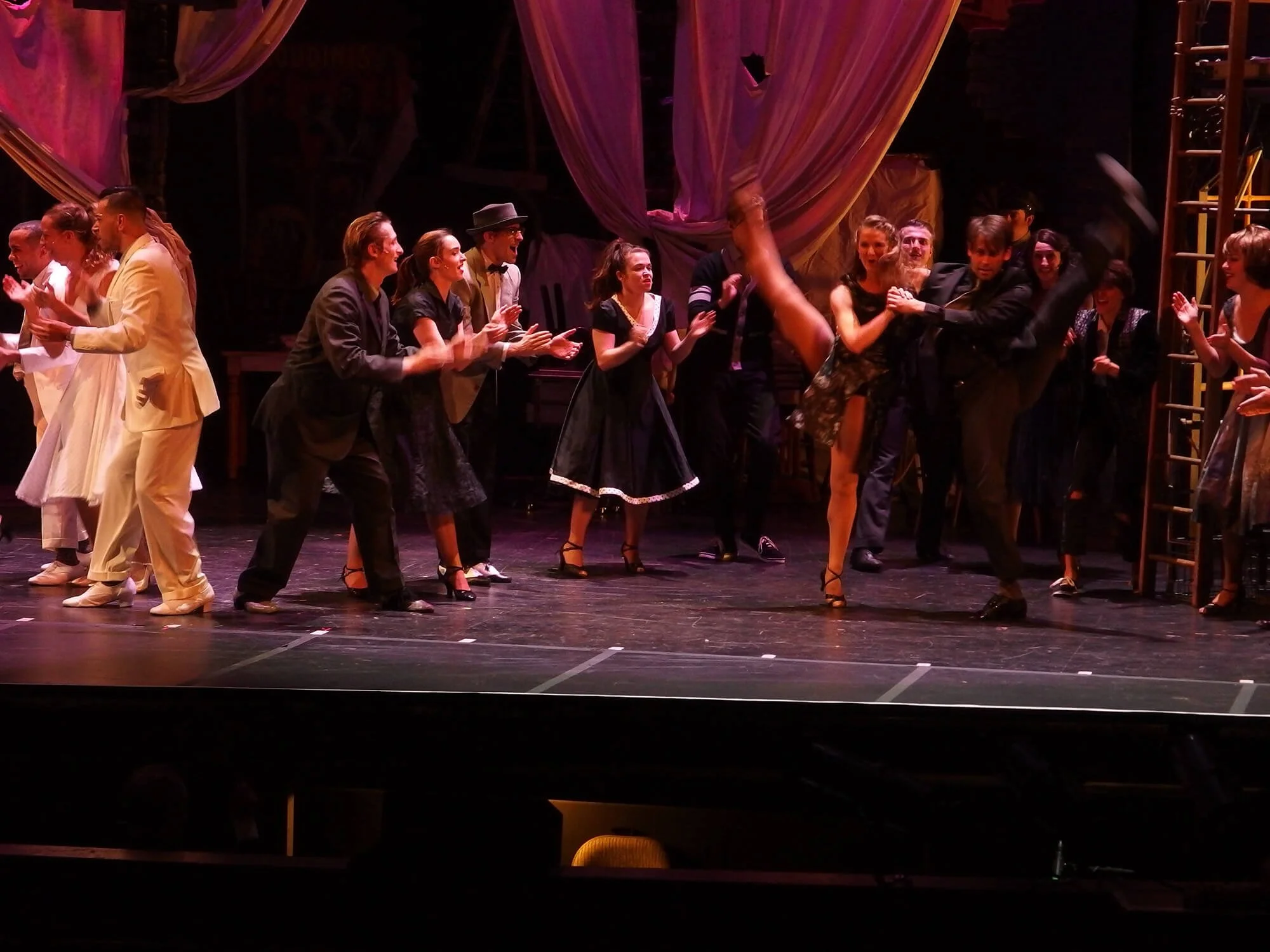 The cast of a stage play bows and applauds together after a performance, with some characters holding hands or embracing on stage.