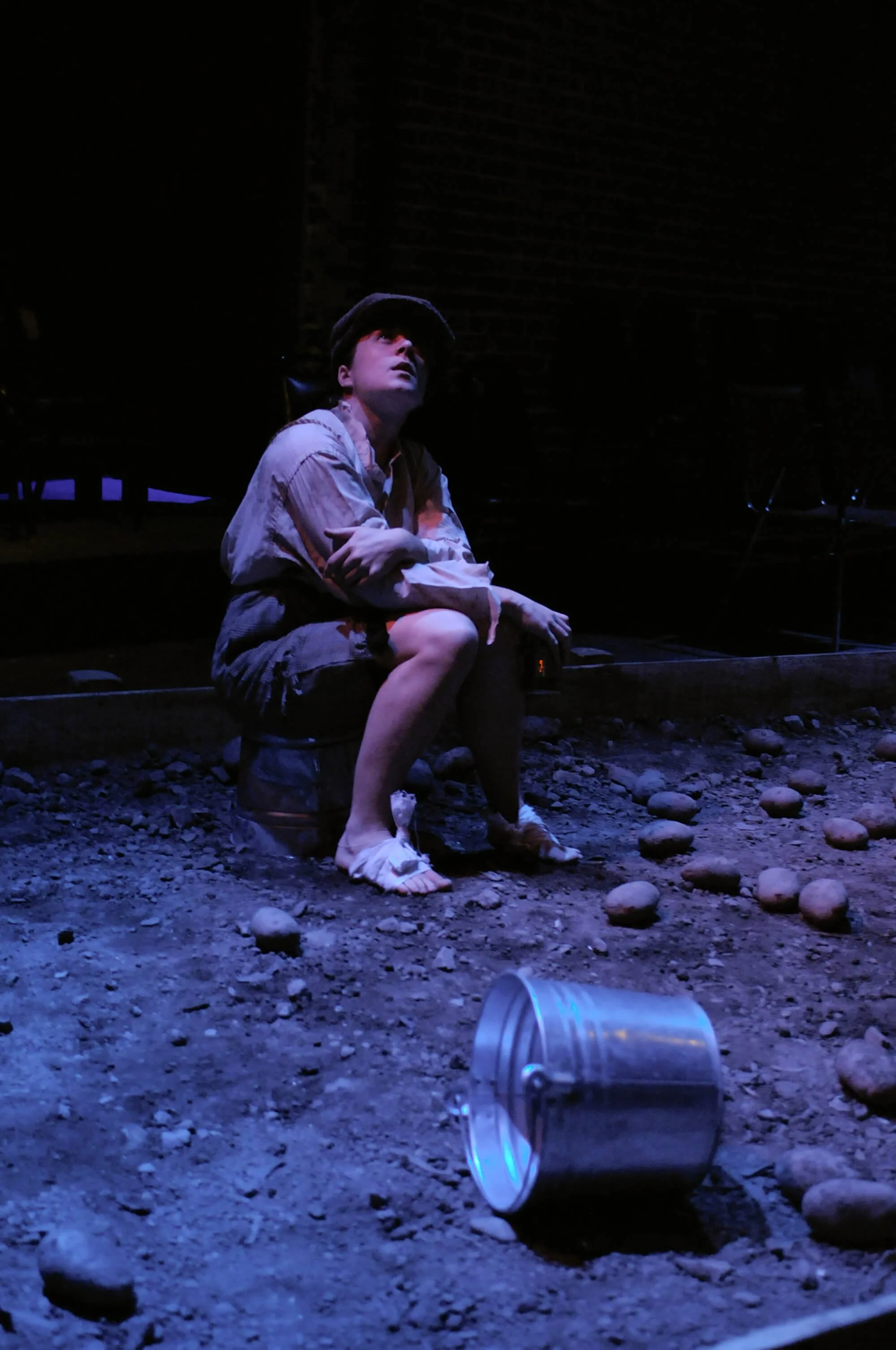A young person with short hair, wearing a cap, sitting on a small stool on a dirt surface, looking upward with a thoughtful expression. They are dressed in a light-colored shirt and shorts. There is a metal can on the ground in front of them and a li