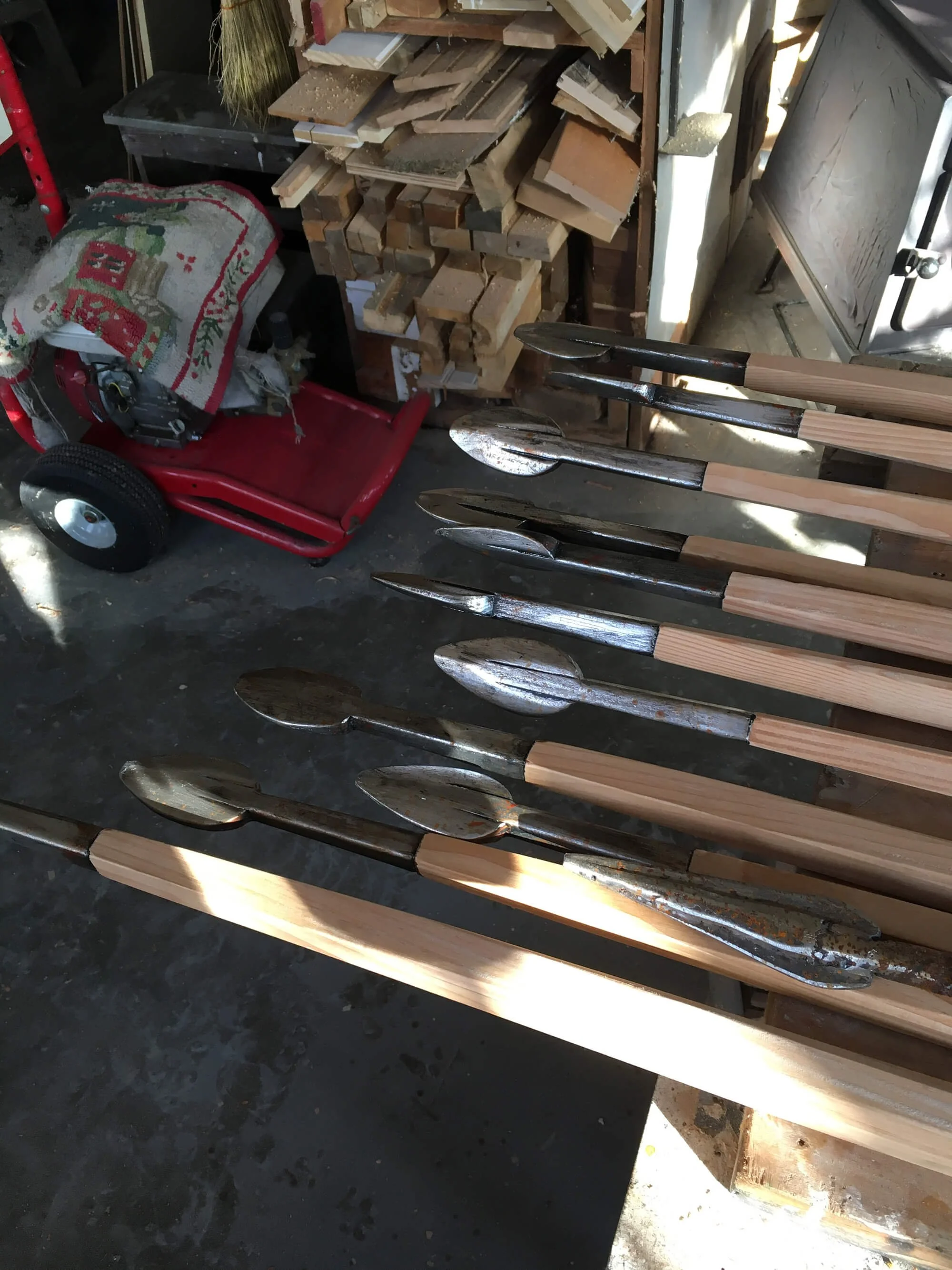 A workbench with several vintage crowbar tools, some with rust, arranged on it. In the background, there are stacked pieces of cut wood and a red wagon with a bag on top in a workshop.