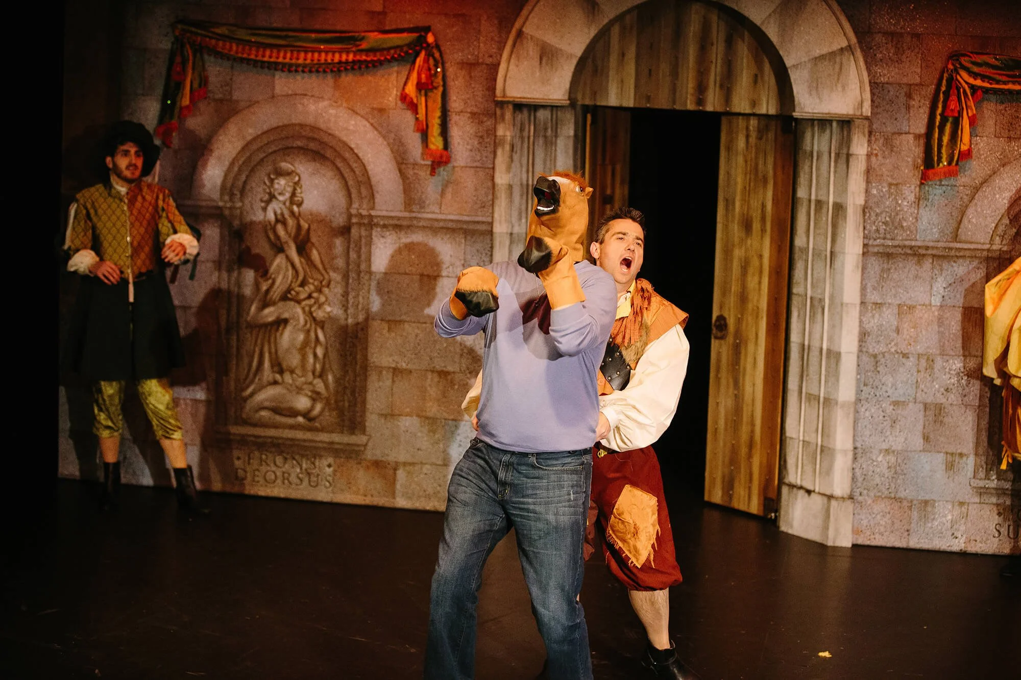 Two men performing on stage, one wearing a horse puppet on his arm, in front of medieval castle-themed backdrop with actors in colorful costumes.