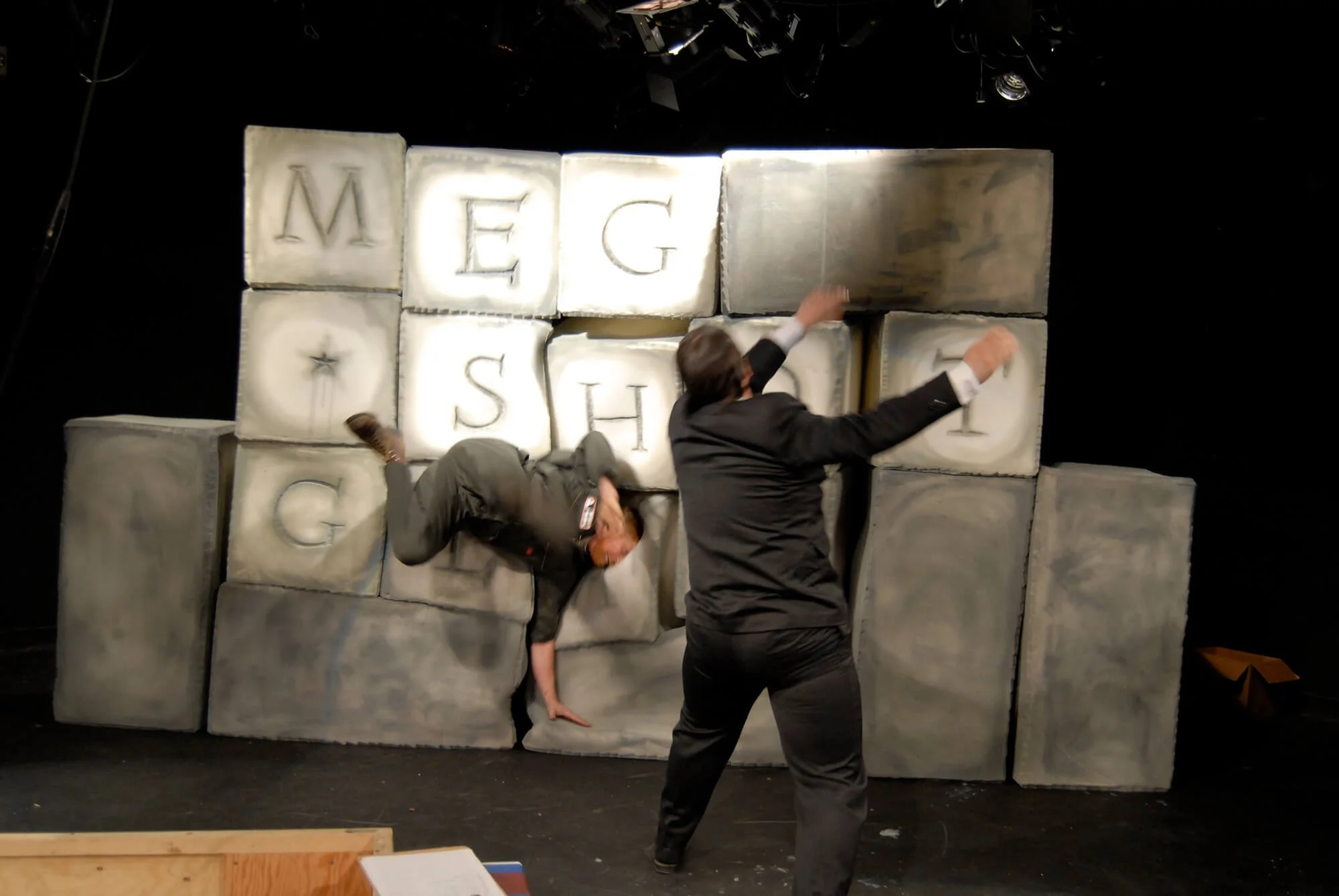 Two people, one in a suit, appear to be falling onto or climbing over a large block wall with letters and symbols. The background shows stage lighting.