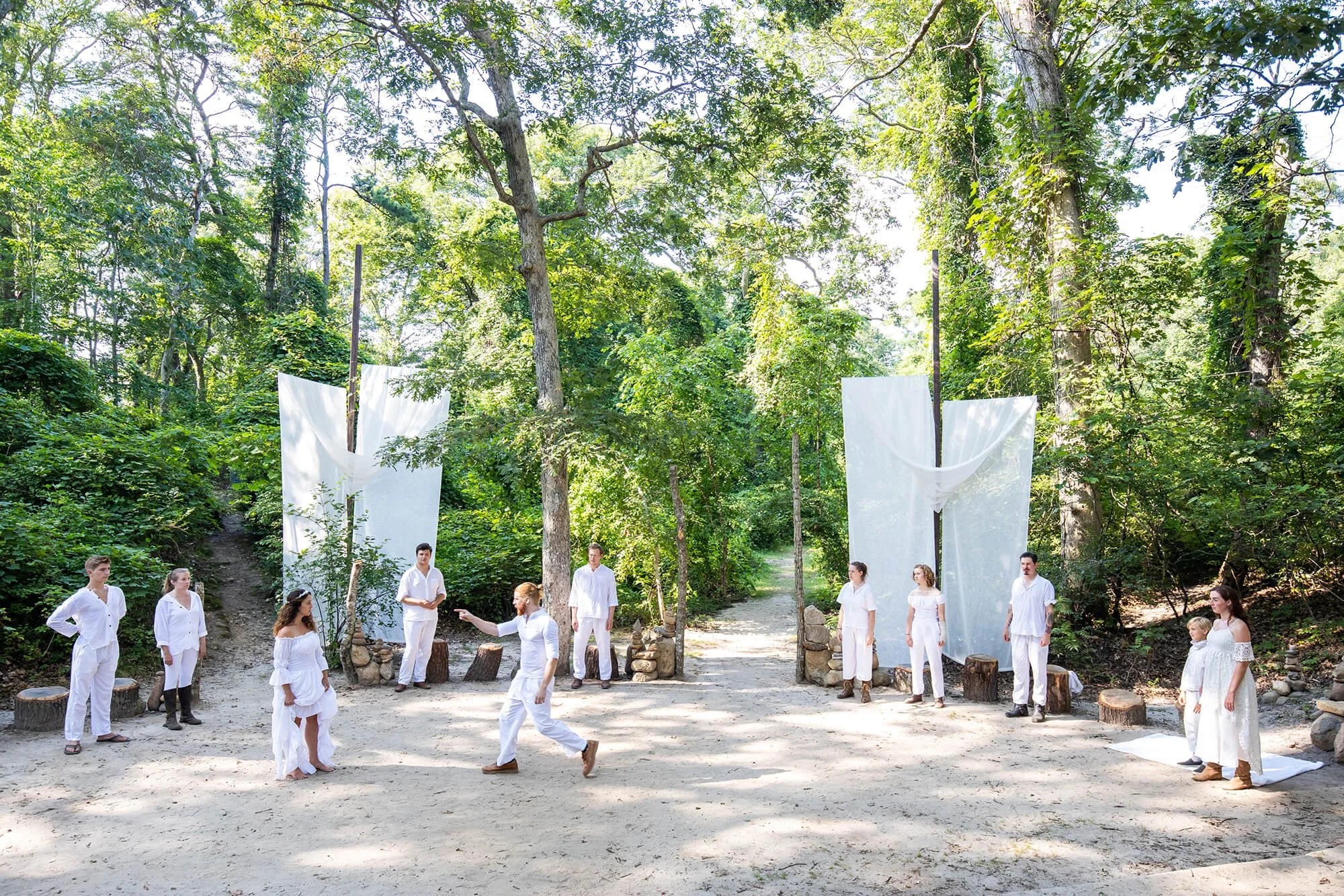 People dressed in white participating in a theatrical performance or ritual in a forest setting, with white fabric panels and natural surroundings.