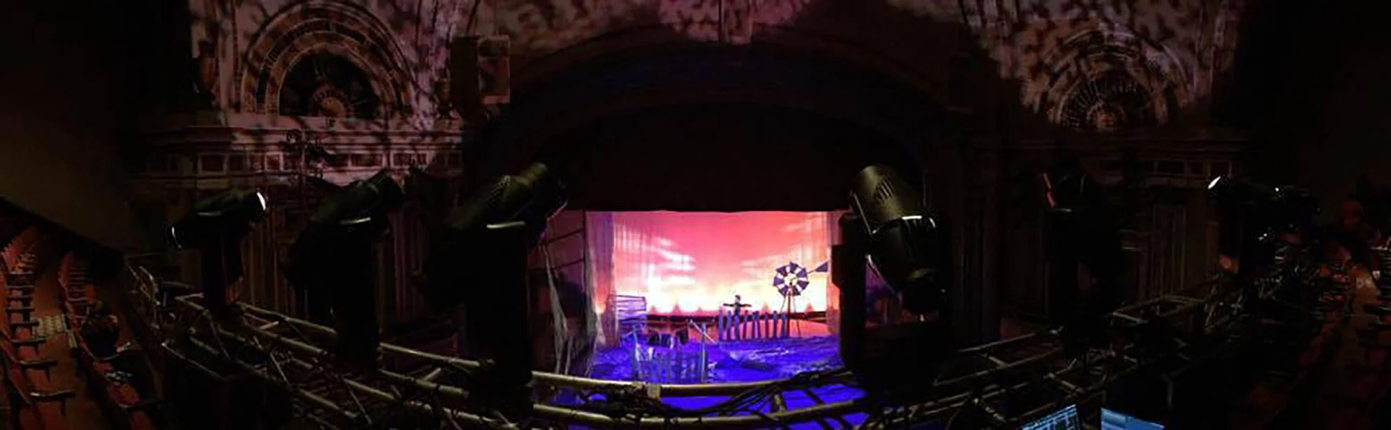 Empty theater with a stage illuminated with colorful lights and a large screen showing a scenic background. Stage lights are set up around the stage, and the theater seats are visible in the foreground.