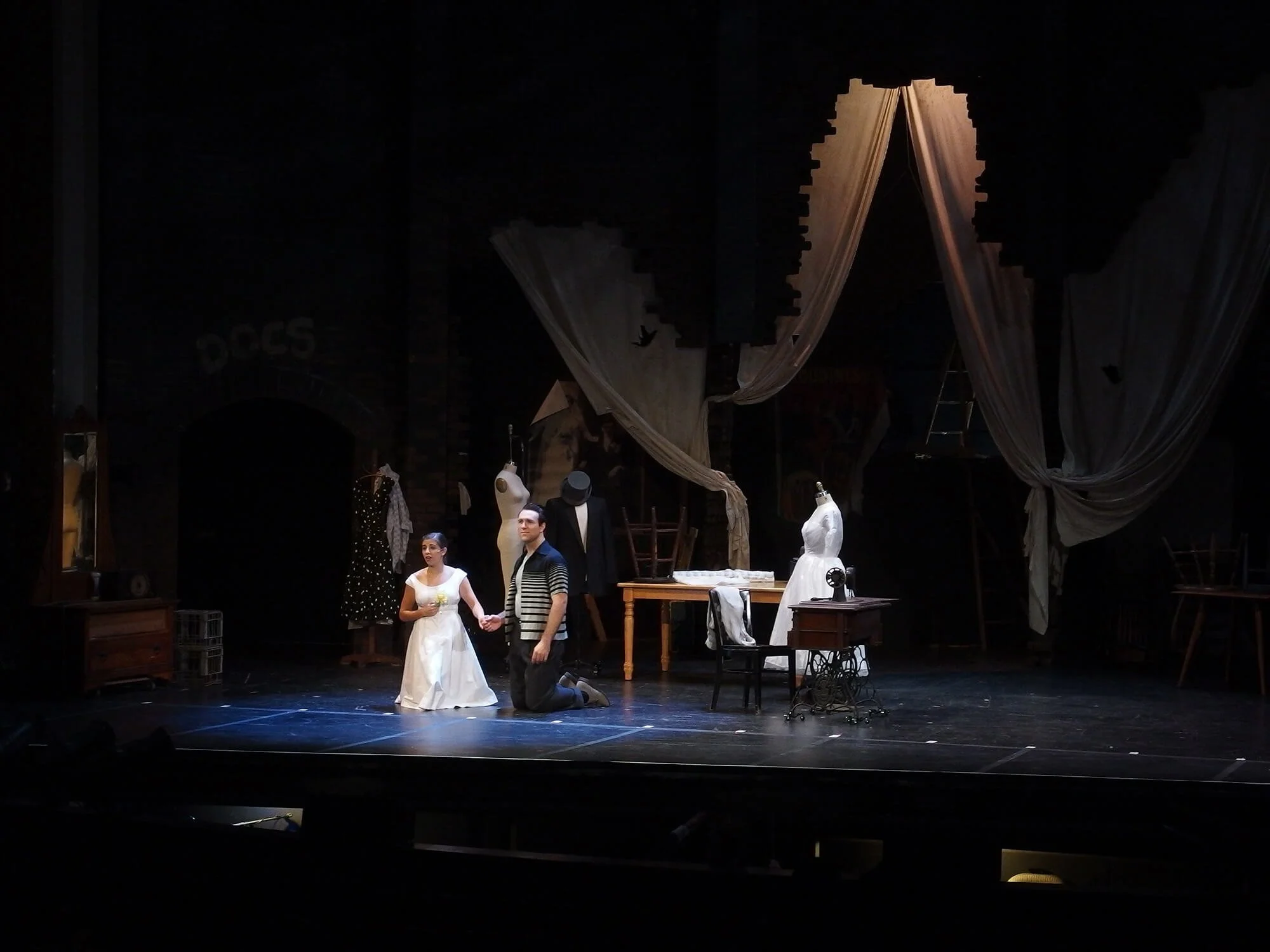 Stage scene from a theater play featuring two actors kneeling, surrounded by mannequins with vintage clothing, antique furniture, and large draped curtains.