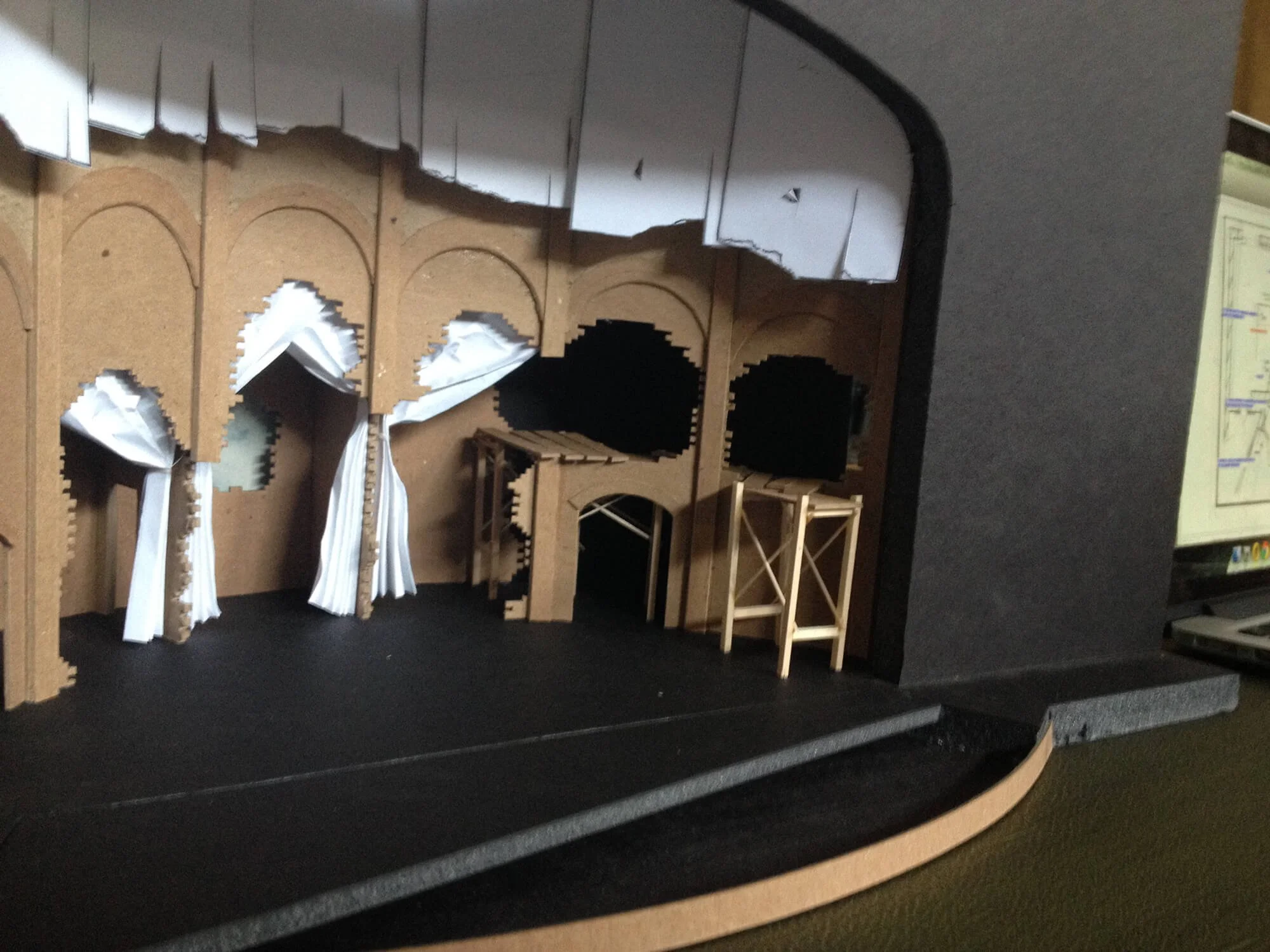 A miniature stage model with wooden arches, curtains, and scaffolding, set against a black background, placed on a black platform with gray edges.